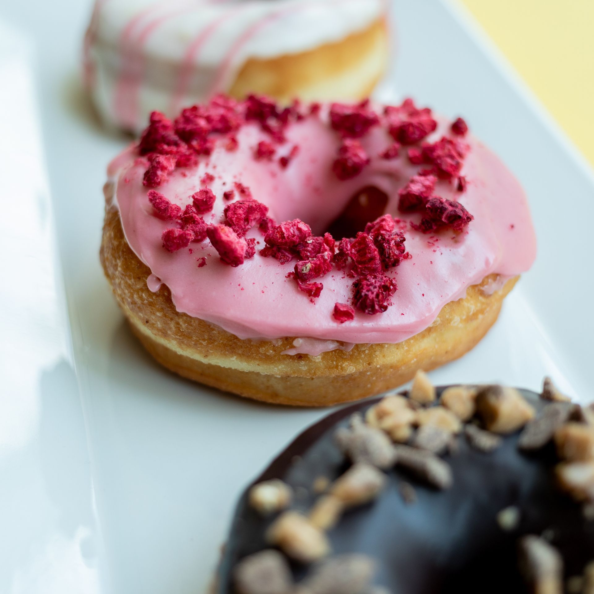 Three frosted donuts on a white rectangular plate. Pink, red and chocolate frosting, with sprinkles.
