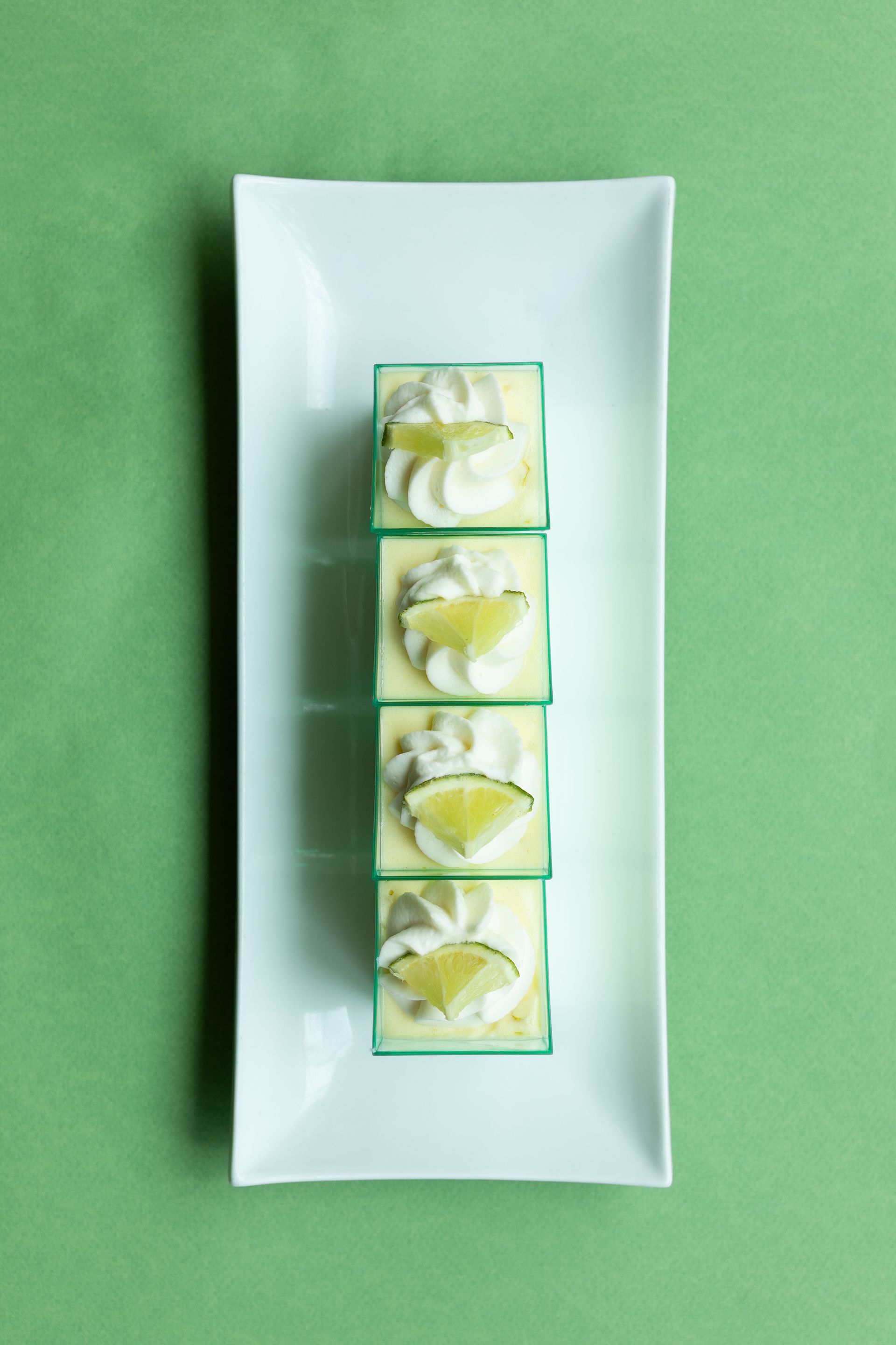Four lime desserts with whipped cream and lime slices on a white rectangular plate. Green background.