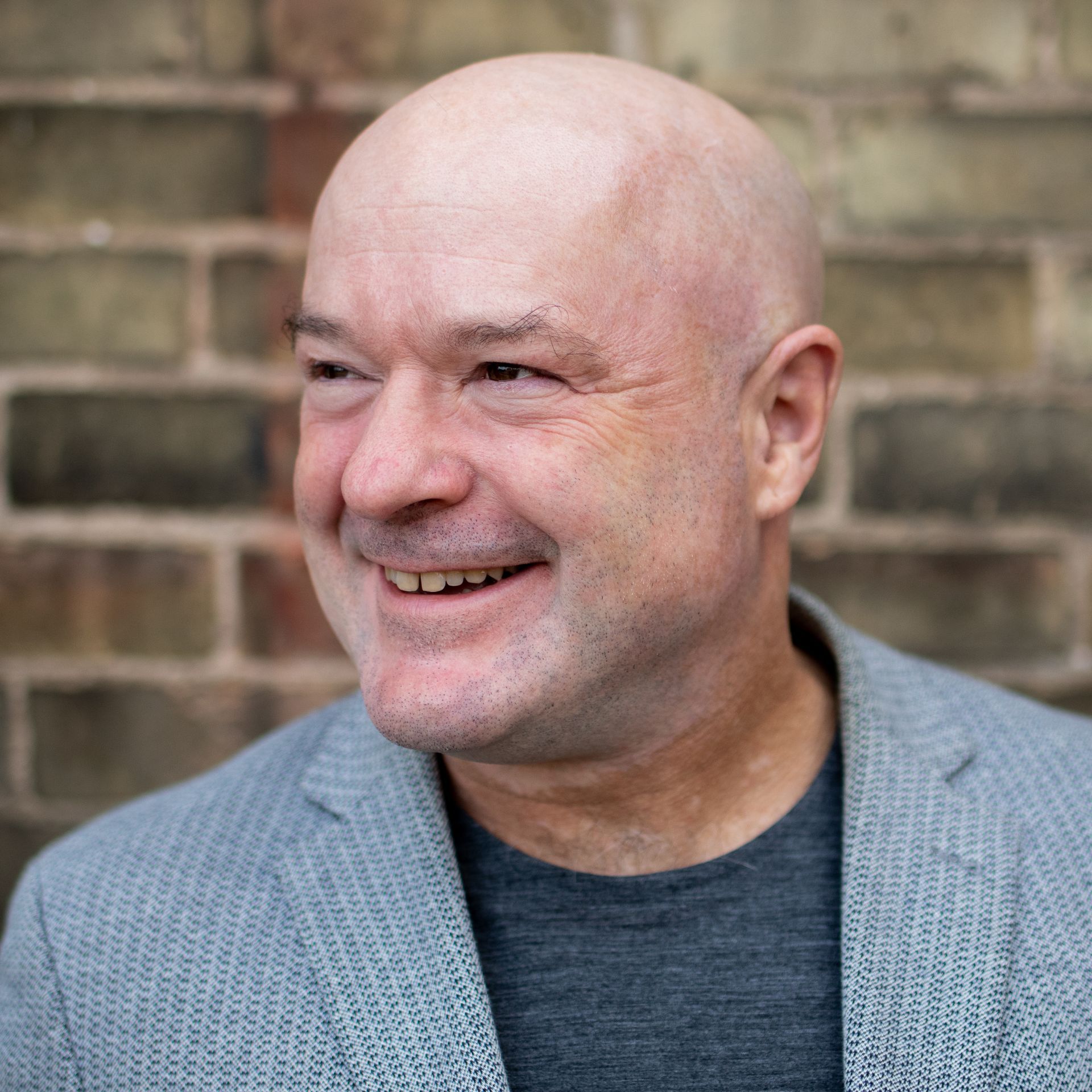 Man with a bald head smiles, looking to the side, wearing a gray blazer and dark gray shirt, against a brick wall.