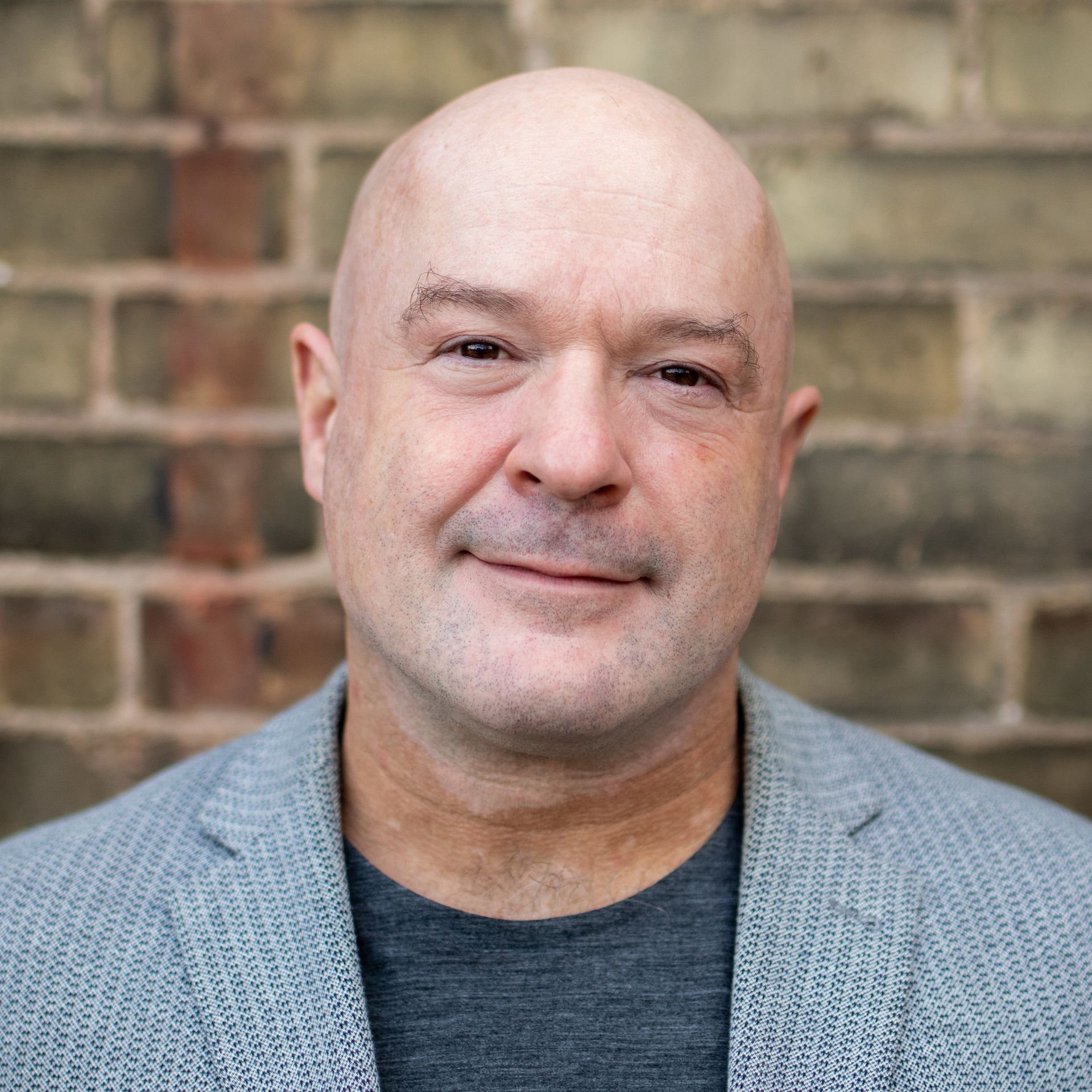 Bald man in gray jacket, smiling in front of a brick wall.