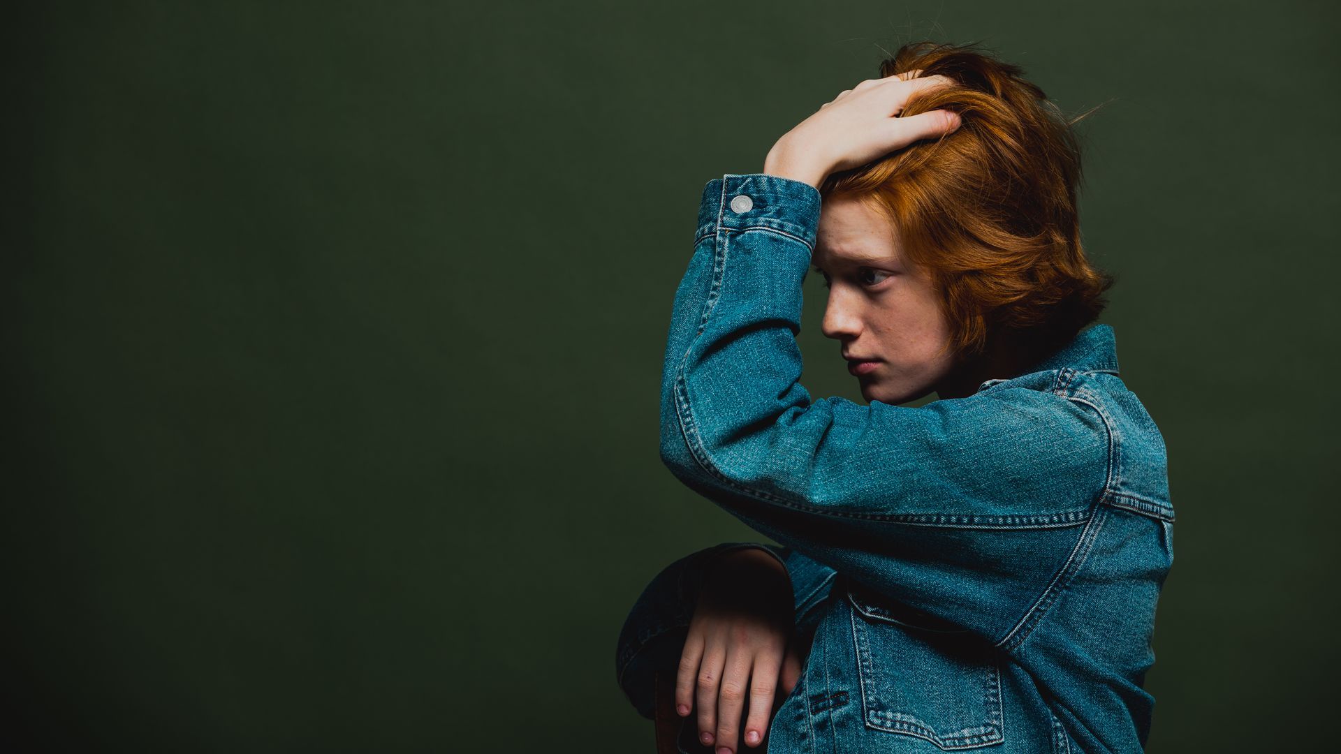 Person with red hair in a denim jacket, hand in hair, against a dark green background.
