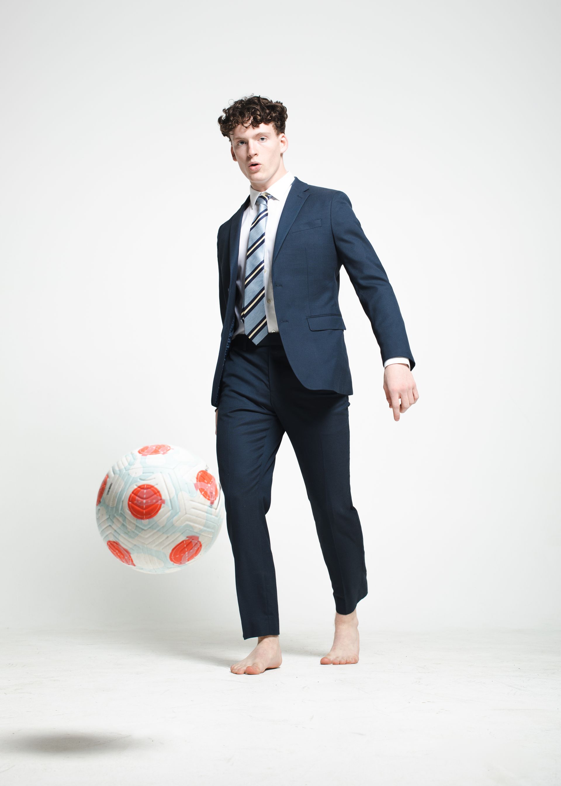 Man in suit barefoot, juggling a large red-and-white soccer ball in a studio.
