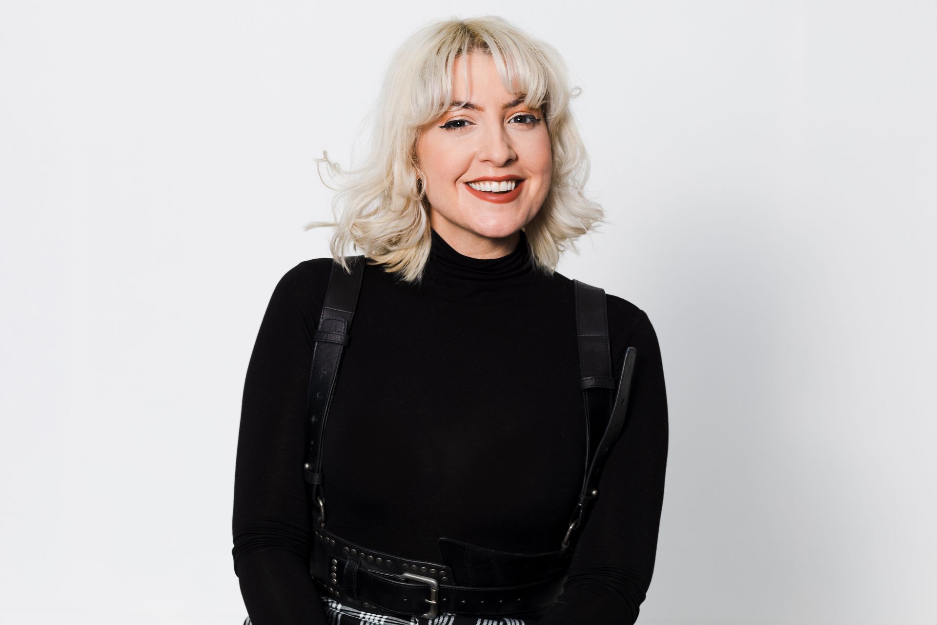 Blonde woman smiles, wearing a black turtleneck and harness, against a white backdrop.