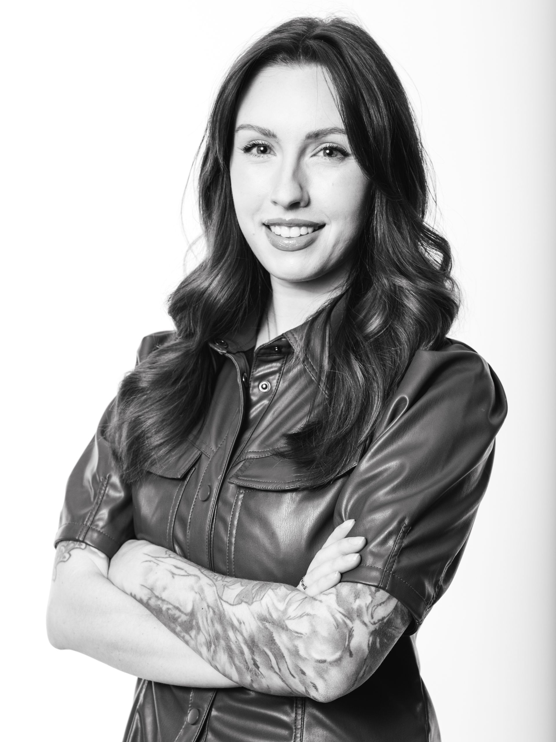 Woman with long, wavy dark hair, arms crossed, wearing a leather-look top and visible arm tattoos, smiling.