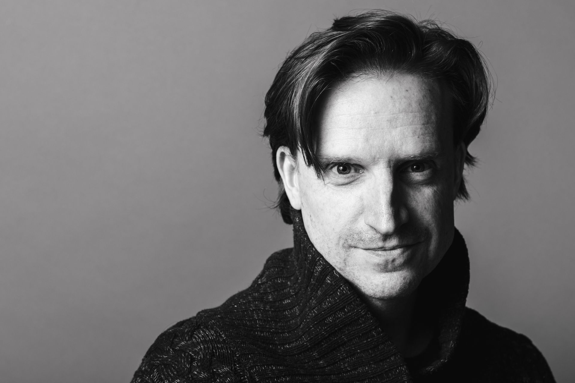 Man with dark hair and a light streak, smiling, wearing a sweater, against a gray background.