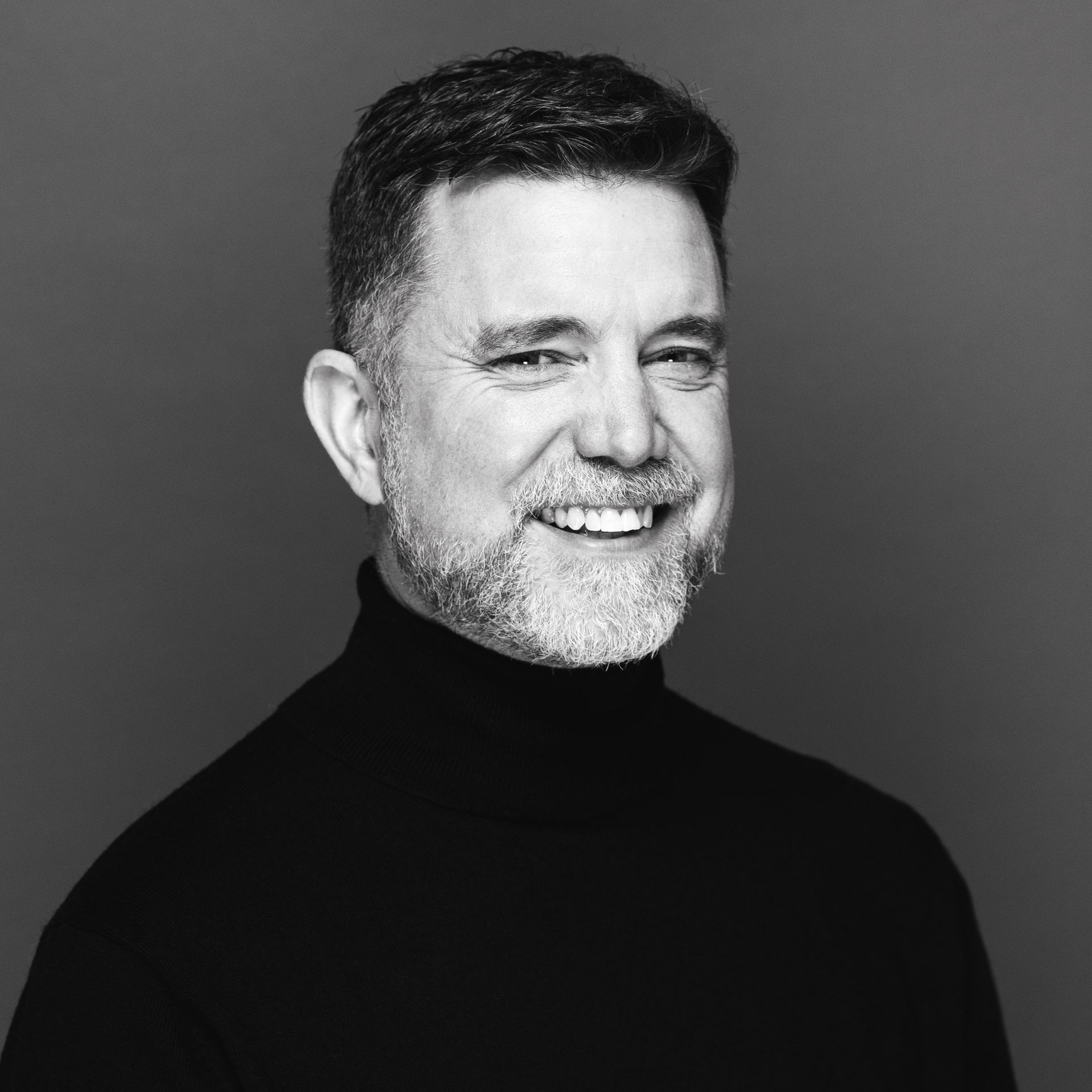 Man with short hair and a beard, wearing a black turtleneck, smiling.