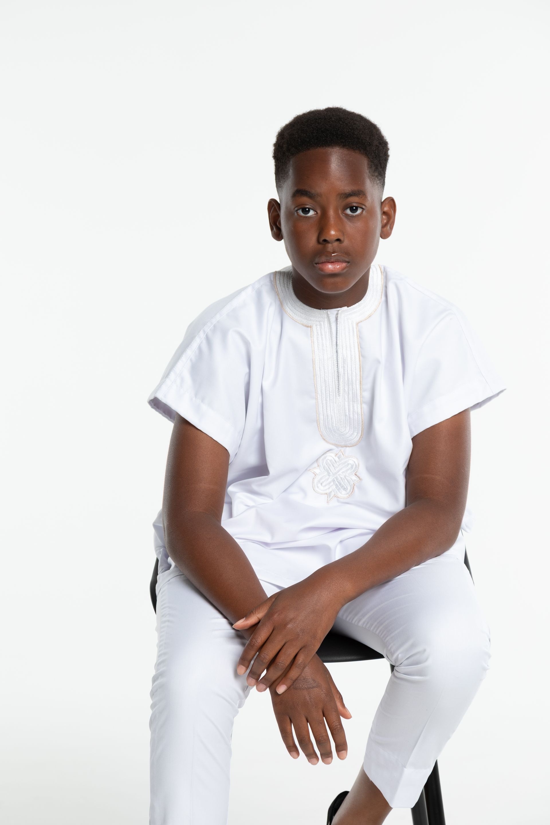 Boy in white traditional clothing, sitting on a stool, looking at the camera against a white backdrop.