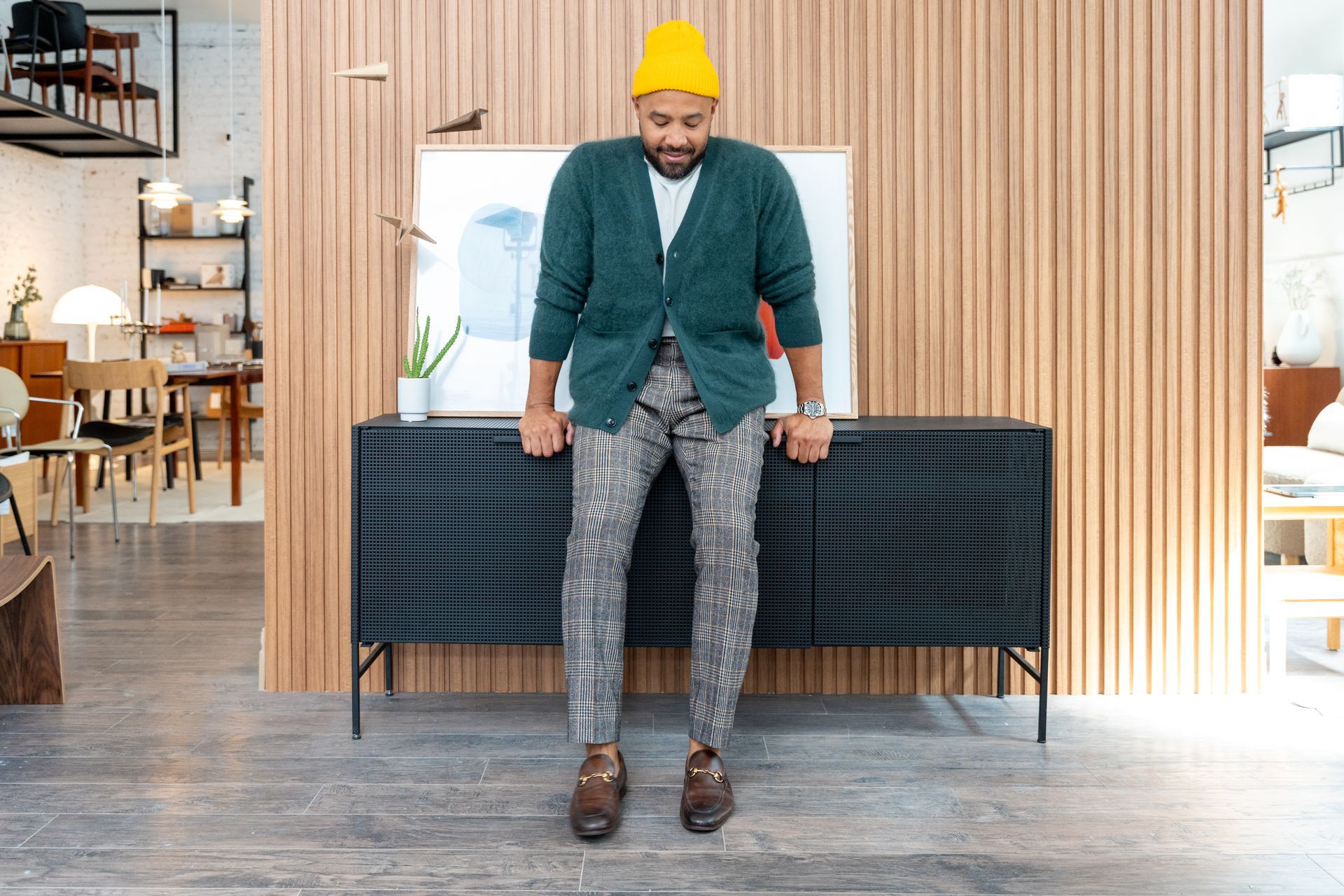 Person in a green cardigan and yellow hat standing in front of a black cabinet.