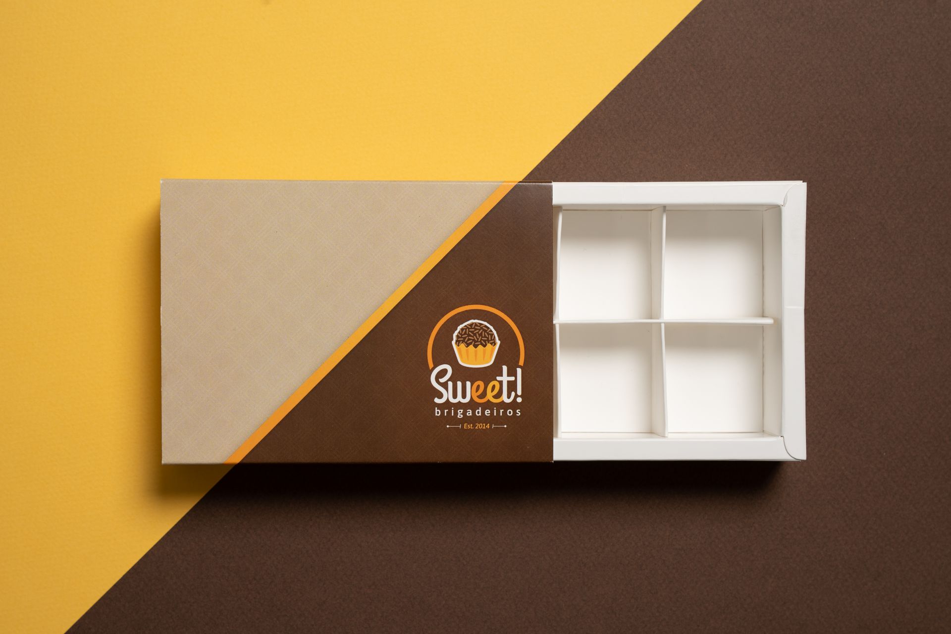 Open, empty chocolate box with logo on brown, angled against yellow and brown background.