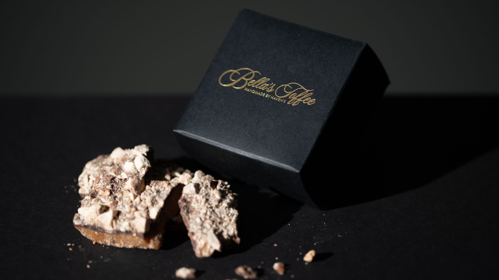 Black gift box with gold logo beside a broken piece of chocolate dessert on a dark surface.