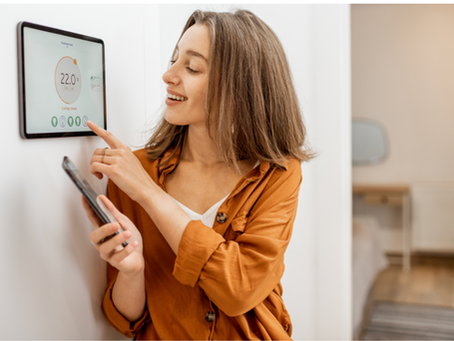Woman smiling at a wall-mounted smart thermostat while holding a smartphone in a bright room