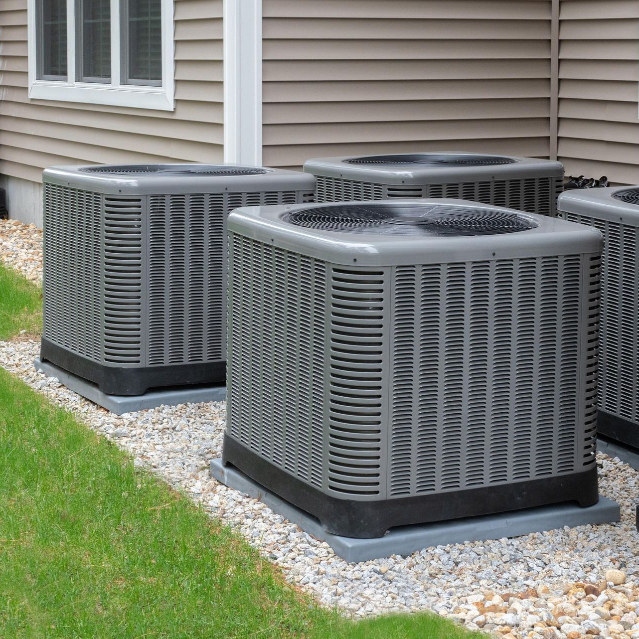 Three gray outdoor air conditioning units beside a house on a gravel bed