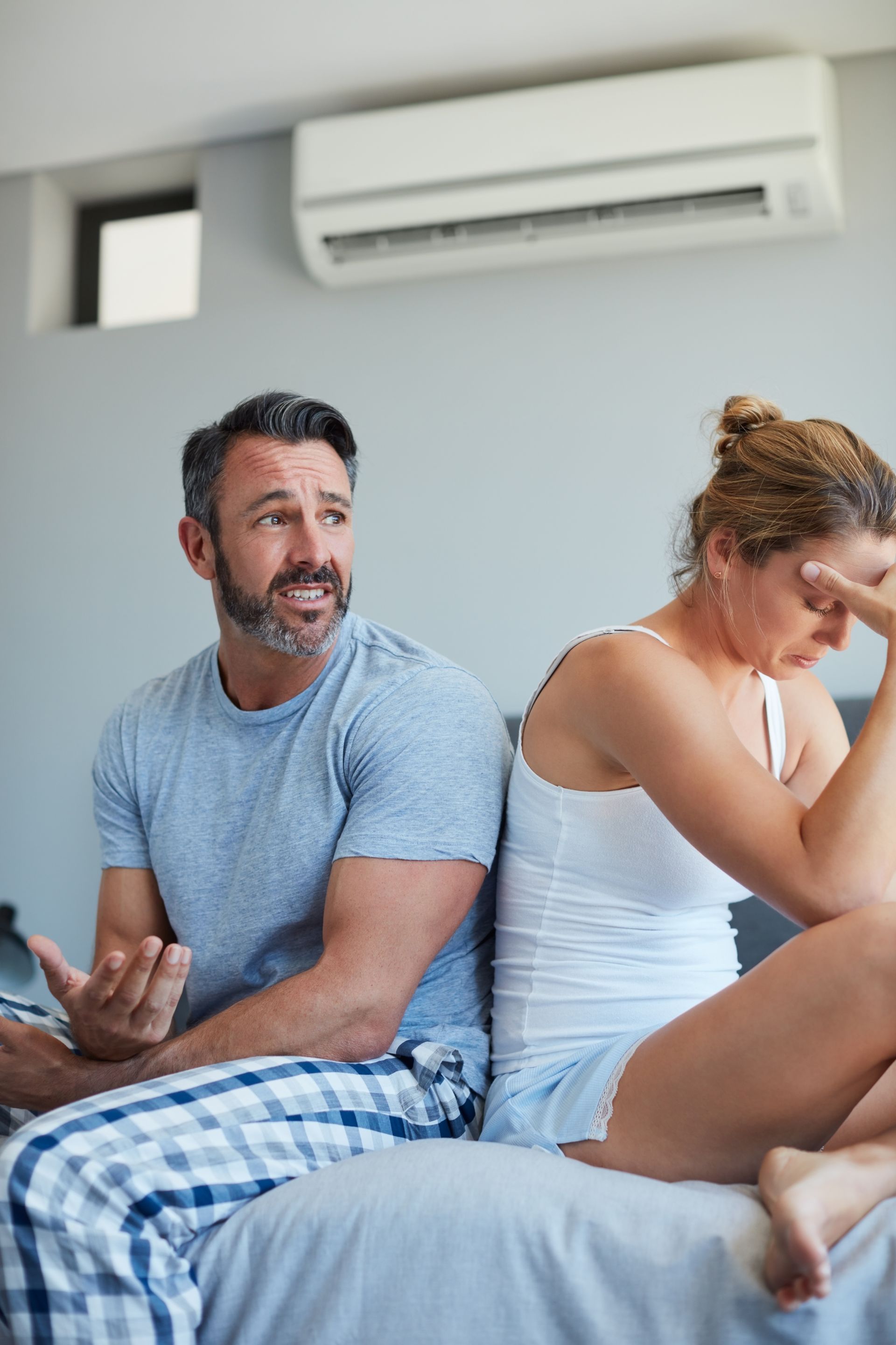 Two people sitting on a bed in a bedroom, one smiling while the other looks upset, with an air conditioner on the wall.