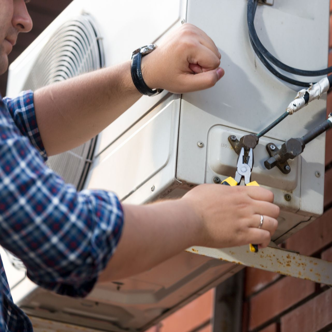 Technician using pliers to repair an outdoor air conditioner unit on a brick wall