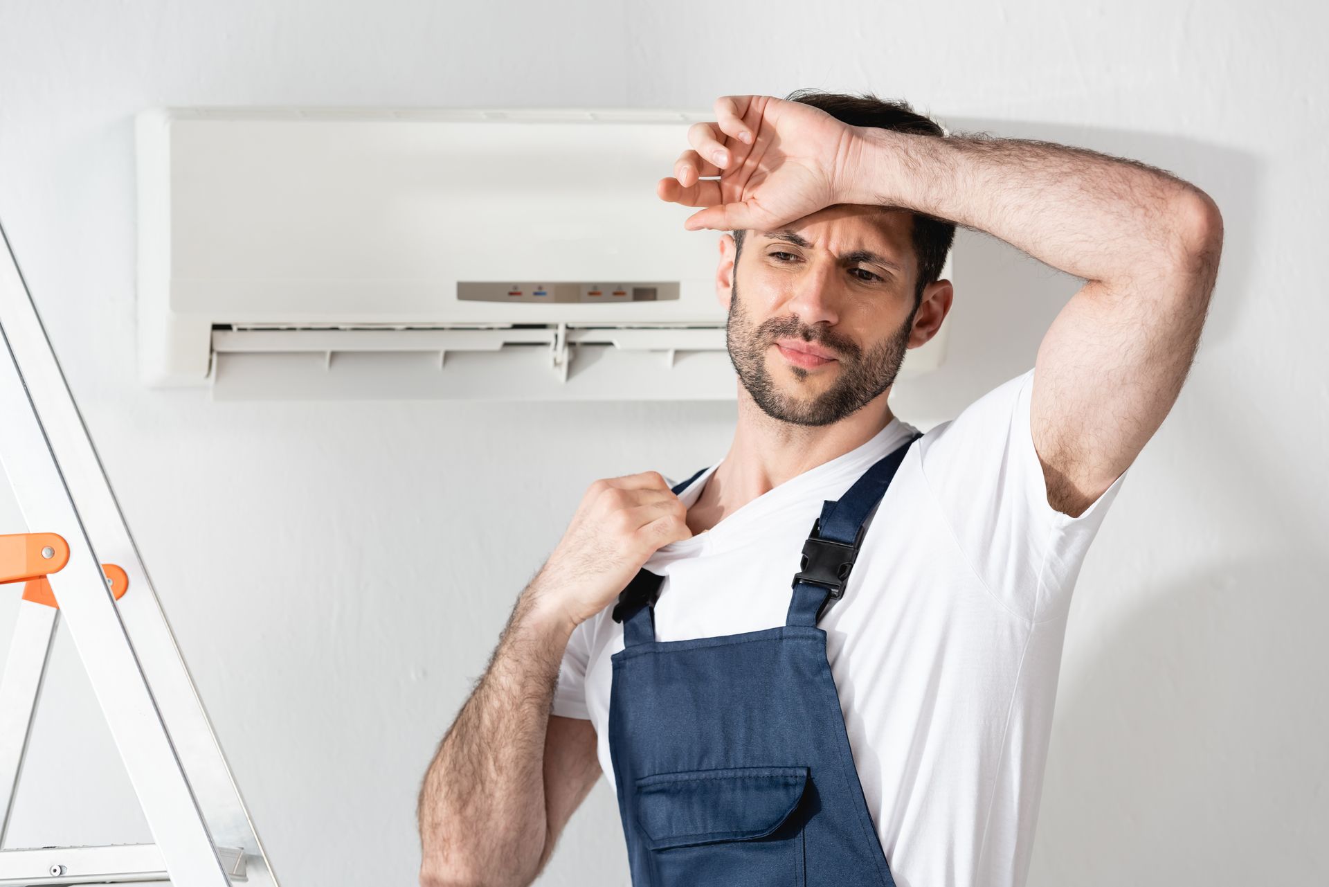 Man sweating and wiping his forehead while standing by a wall-mounted air conditioner in a home interior