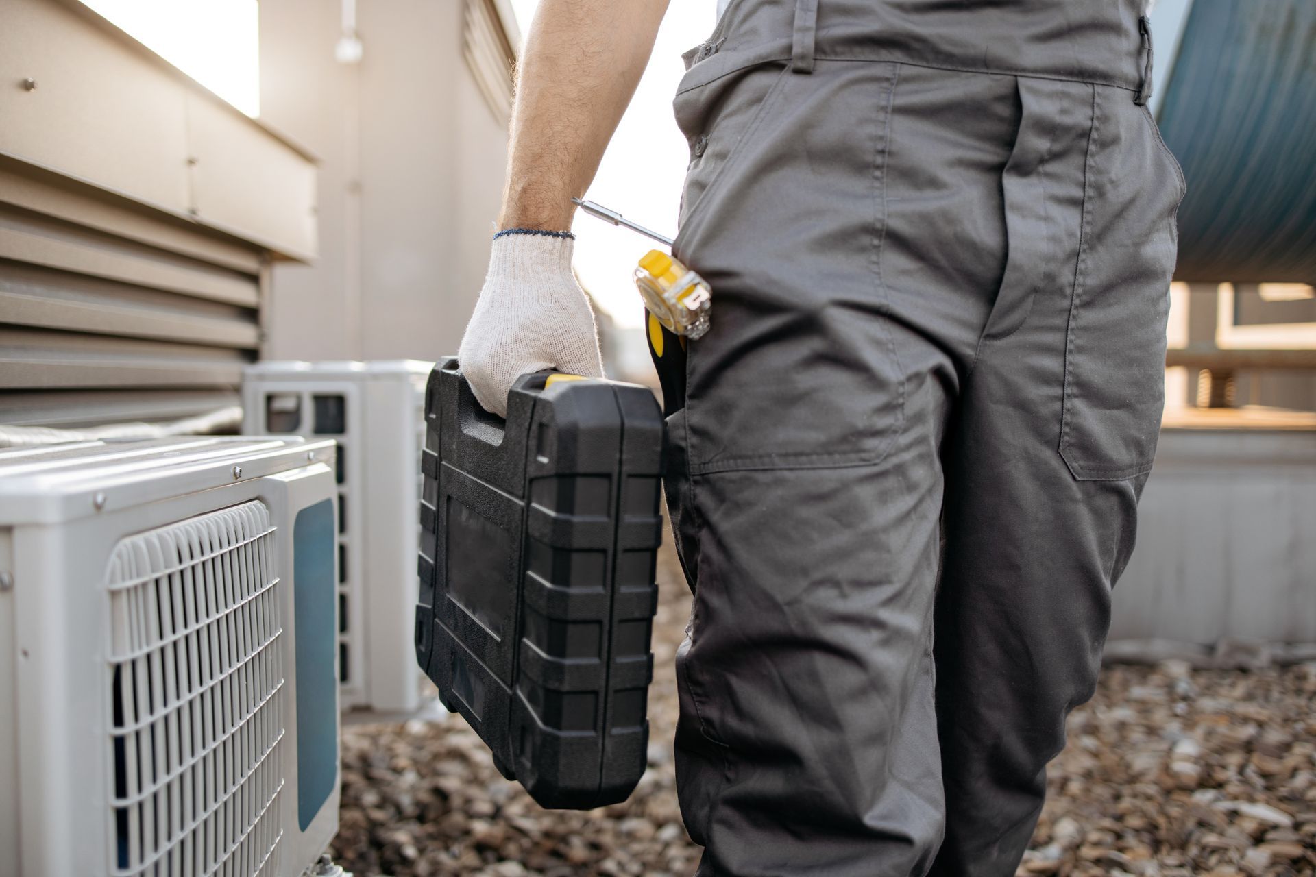 Technician carrying a black toolbox beside outdoor HVAC units on gravel.