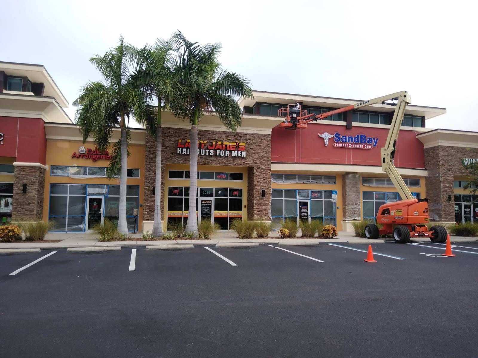 Strip mall storefront with palm trees, a ladder lift, and an empty parking lot.