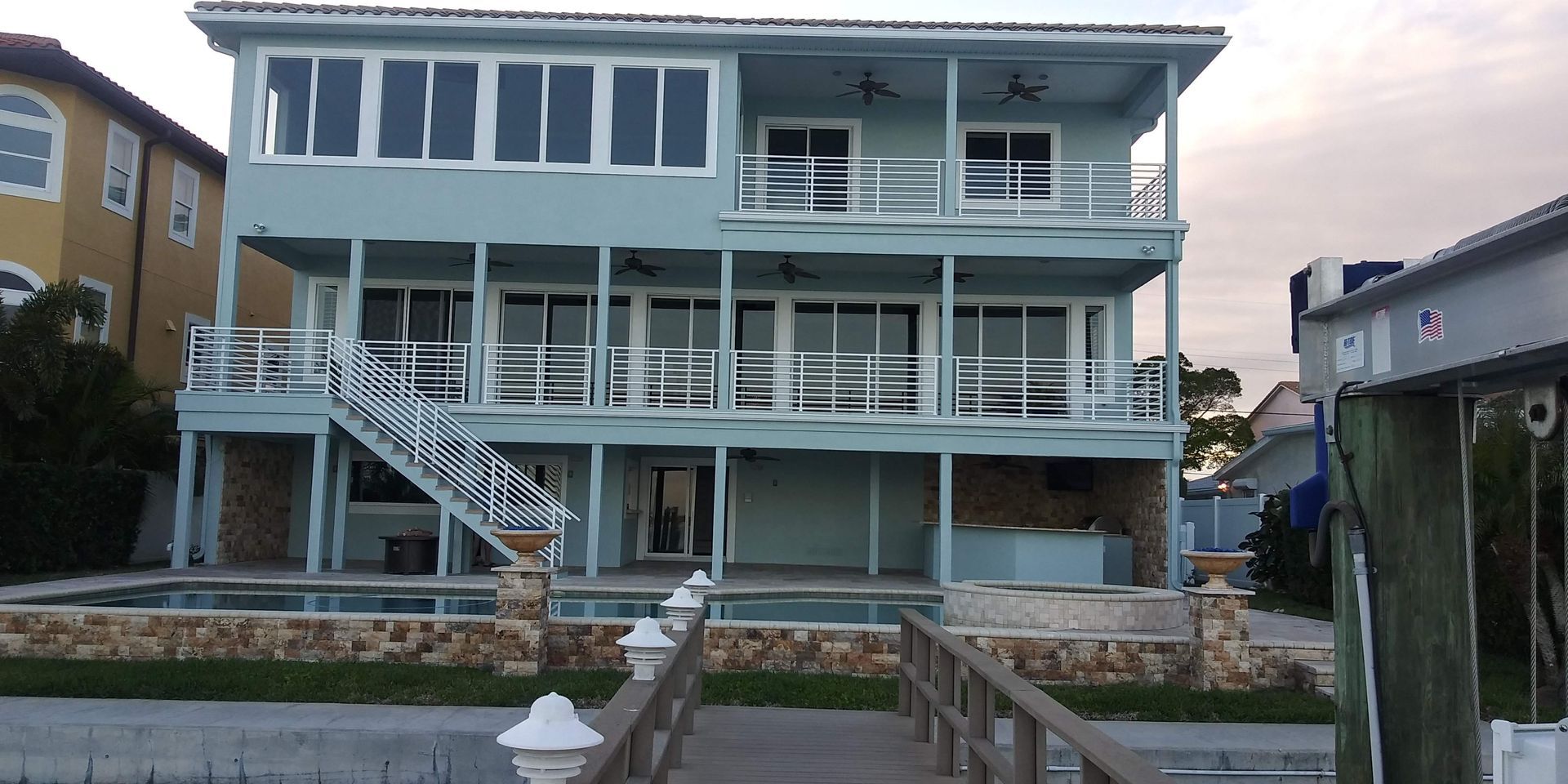 Blue-gray two-story beach house with wraparound porch and exterior stairs behind a stone wall
