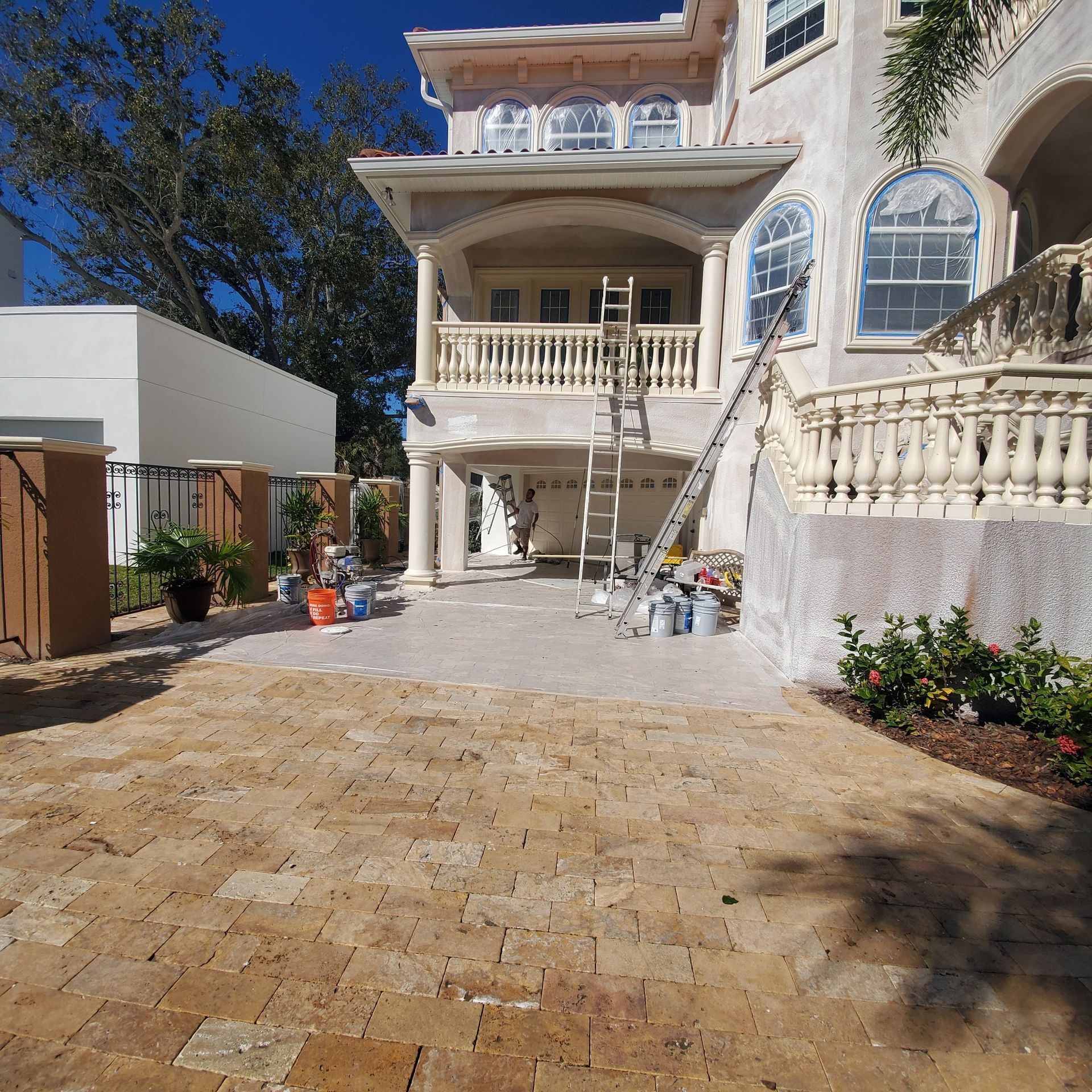 Mansion courtyard with stone driveway, white balcony, ladder, and building supplies under a bright blue sky