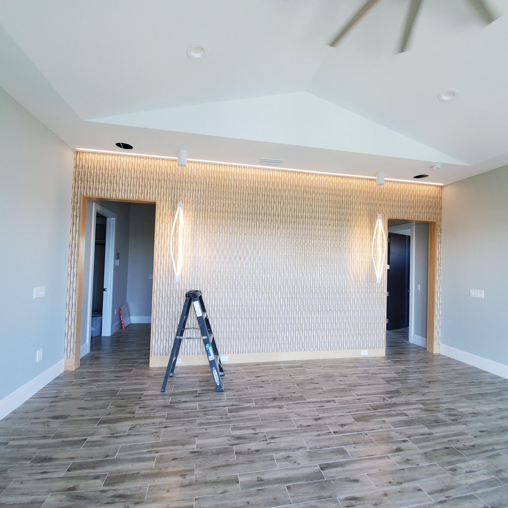 Empty modern living room with gray walls, wood-look floor, vaulted ceiling, and a ladder in front of a patterned accent wall