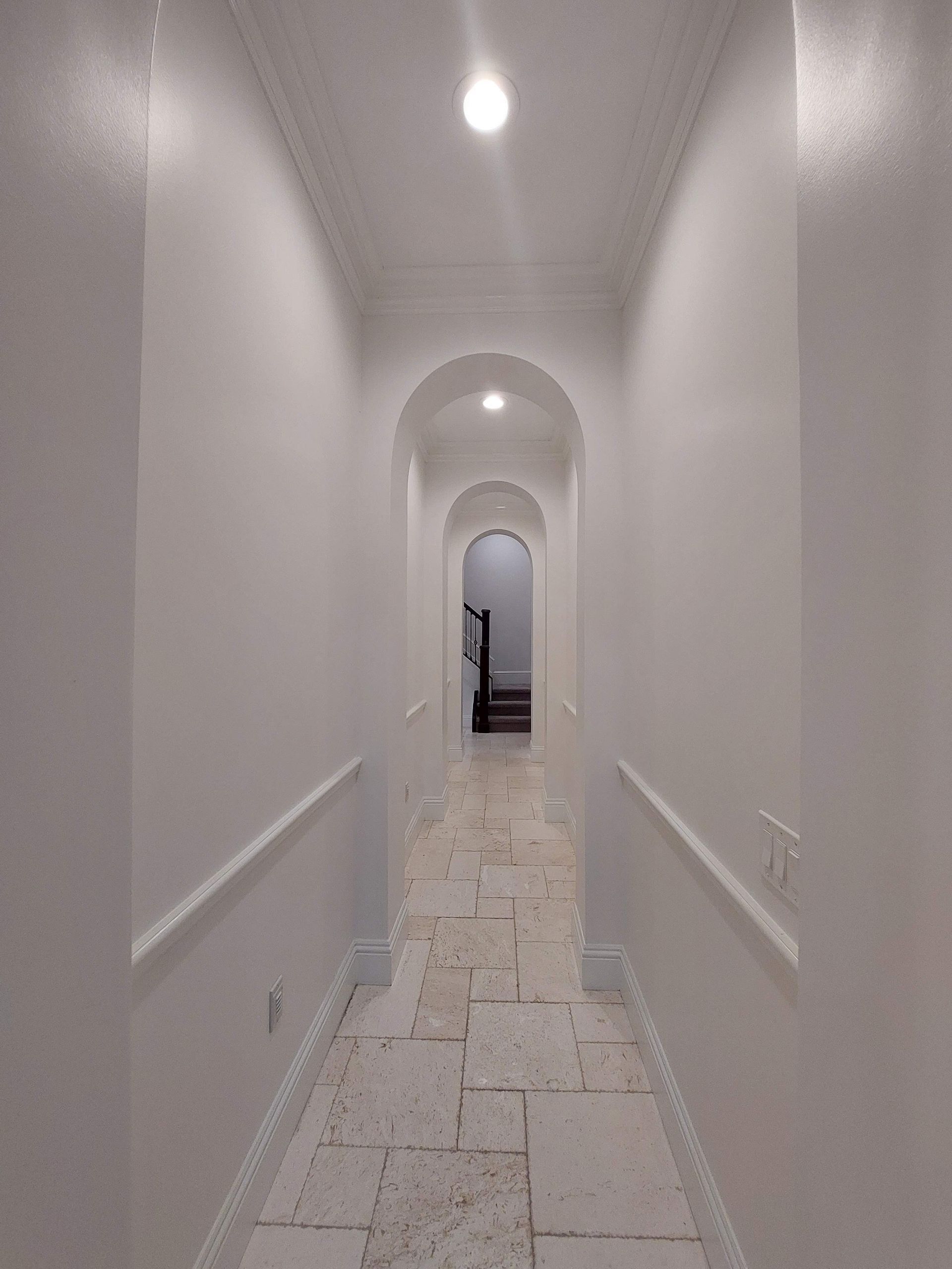 Long white hallway with arched doorways, wall sconces, and tiled floor leading to a distant open door