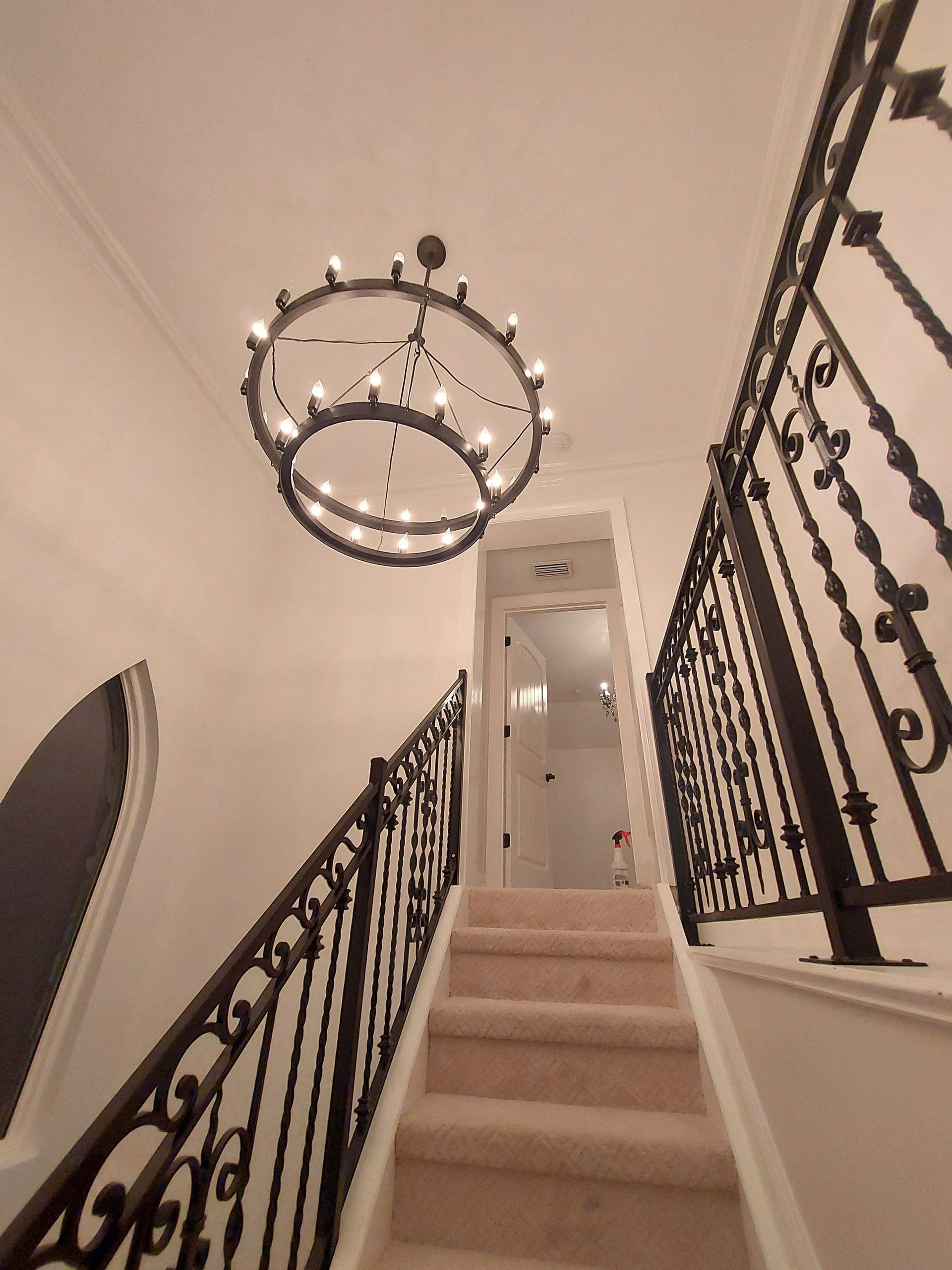 Carpeted staircase with black wrought-iron railings and a chandelier above, leading to an upstairs hallway.