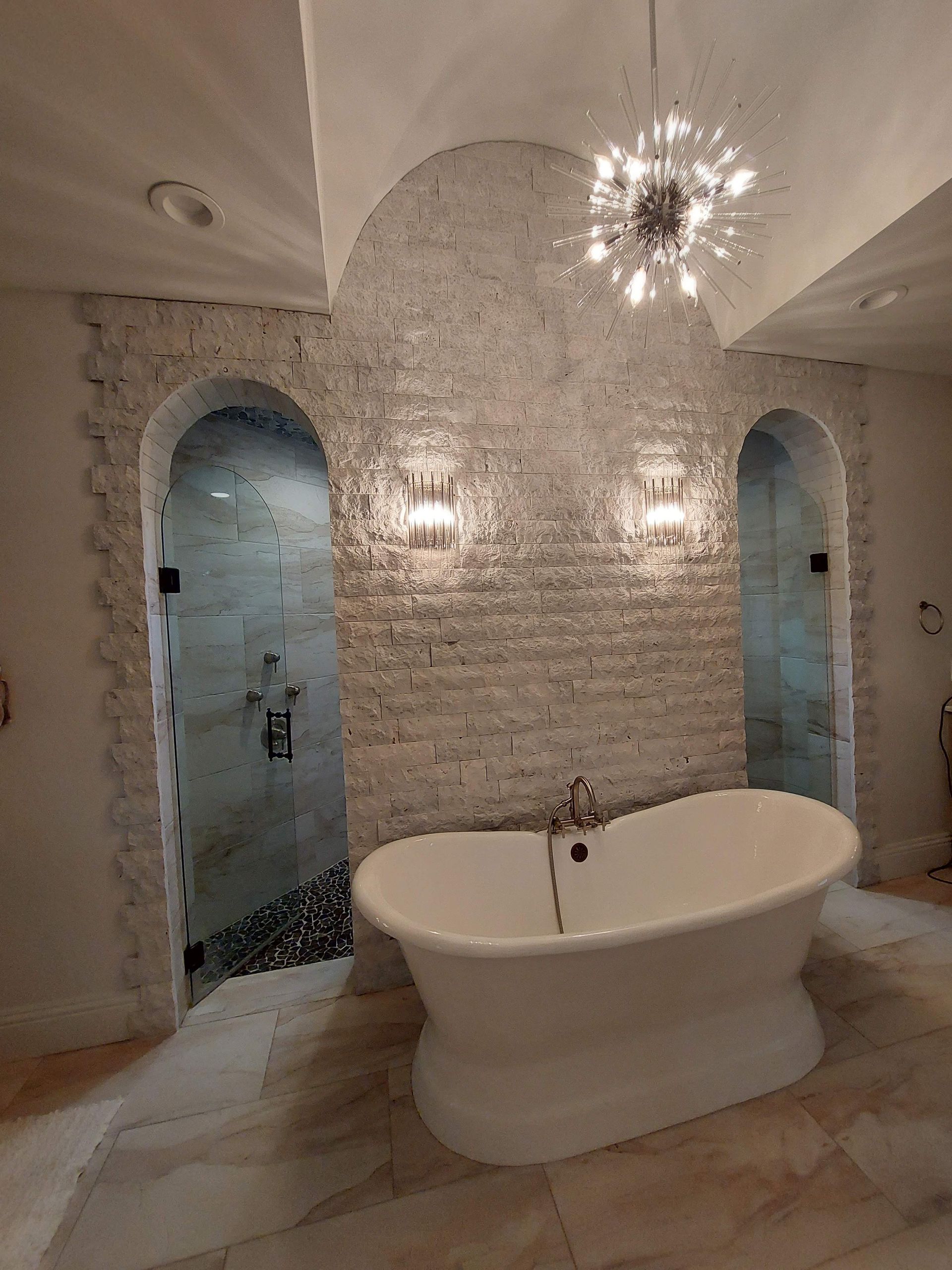 Elegant white marble bathroom with freestanding tub, arched doorways, and chandelier lighting