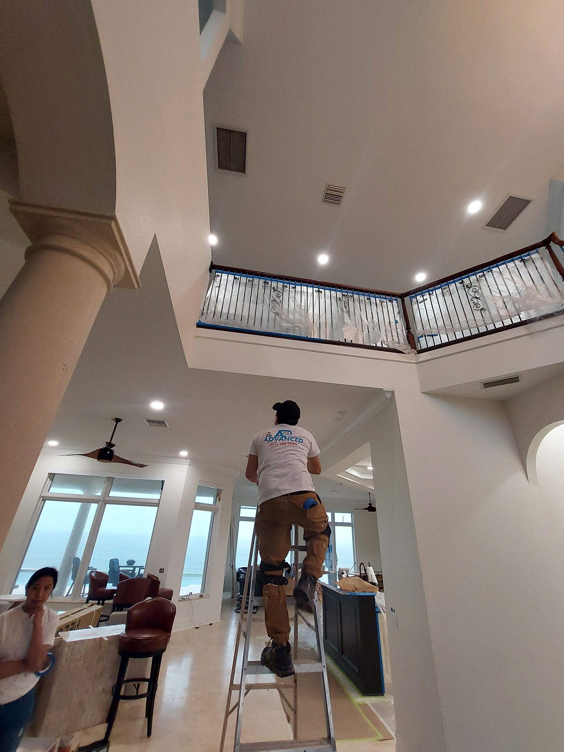 Person stands on a ladder in a bright, two-story room with arched openings and a balcony railing.