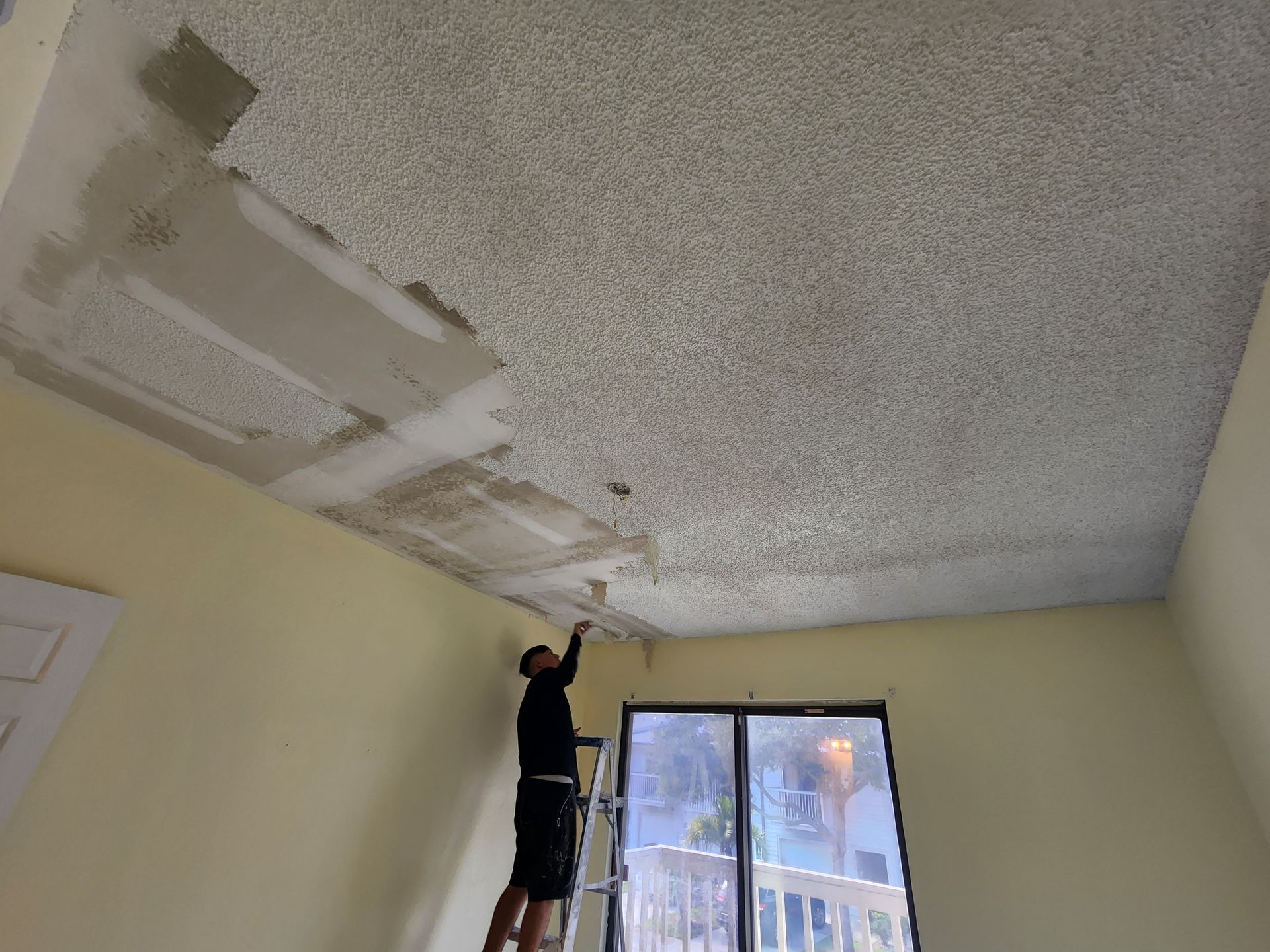 Person on a ladder patching a damaged ceiling in a room with a window. Person on a ladder patching a damaged ceiling in a room with a window.