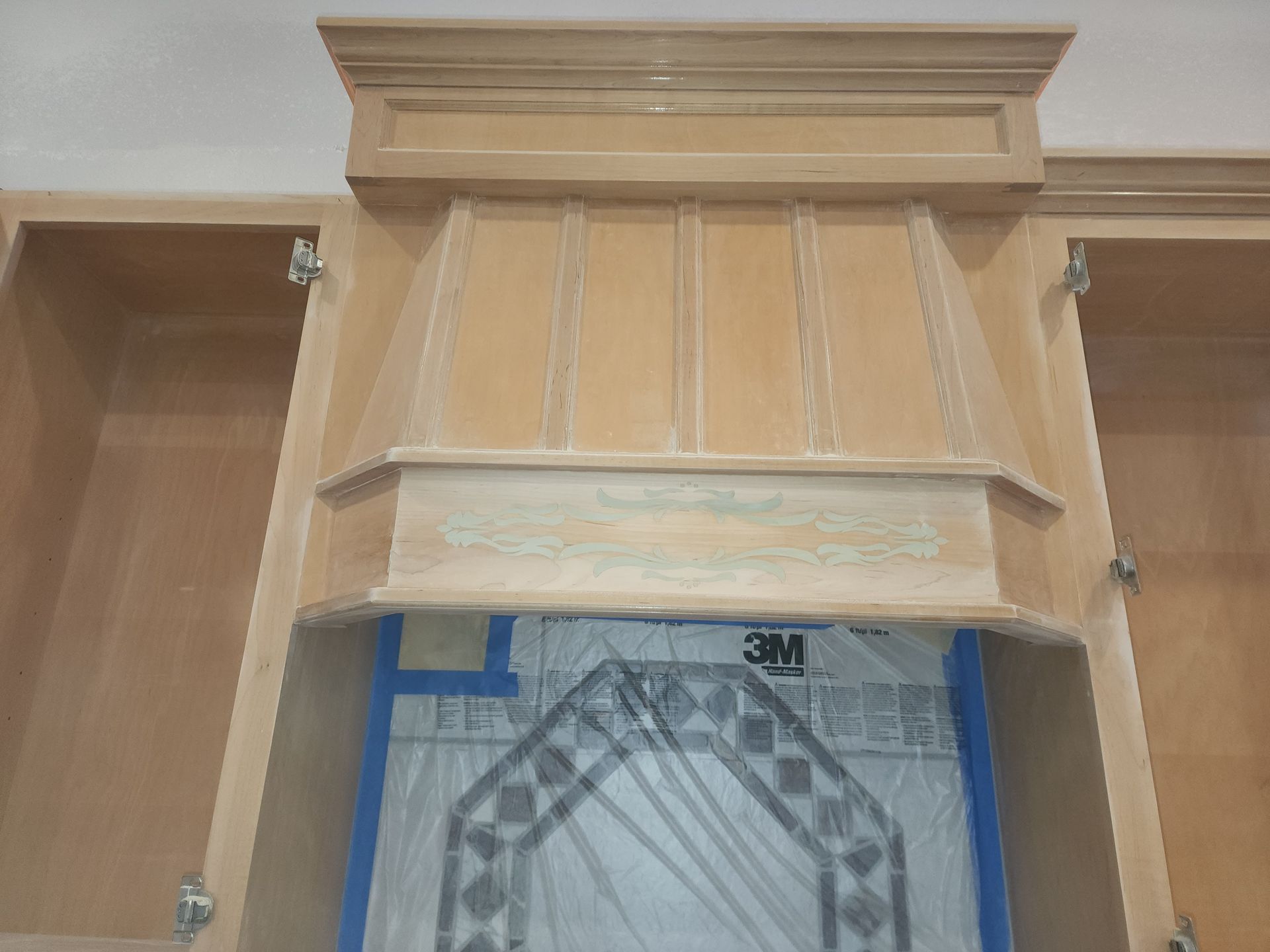 Wooden wall-mounted cabinet with a light tan finish and blue protective tape around the opening.