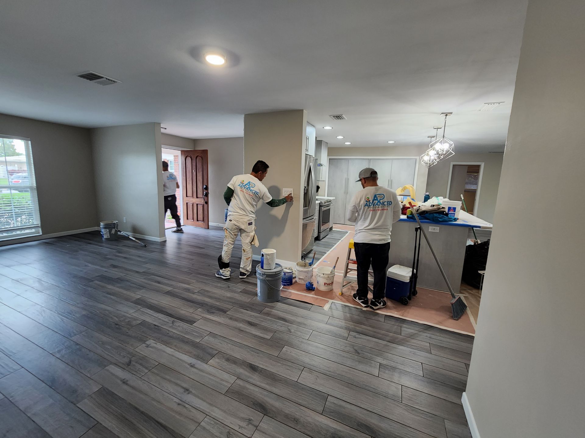 Workers painting a room with dark wood floors and gray walls in a bright, empty interior.