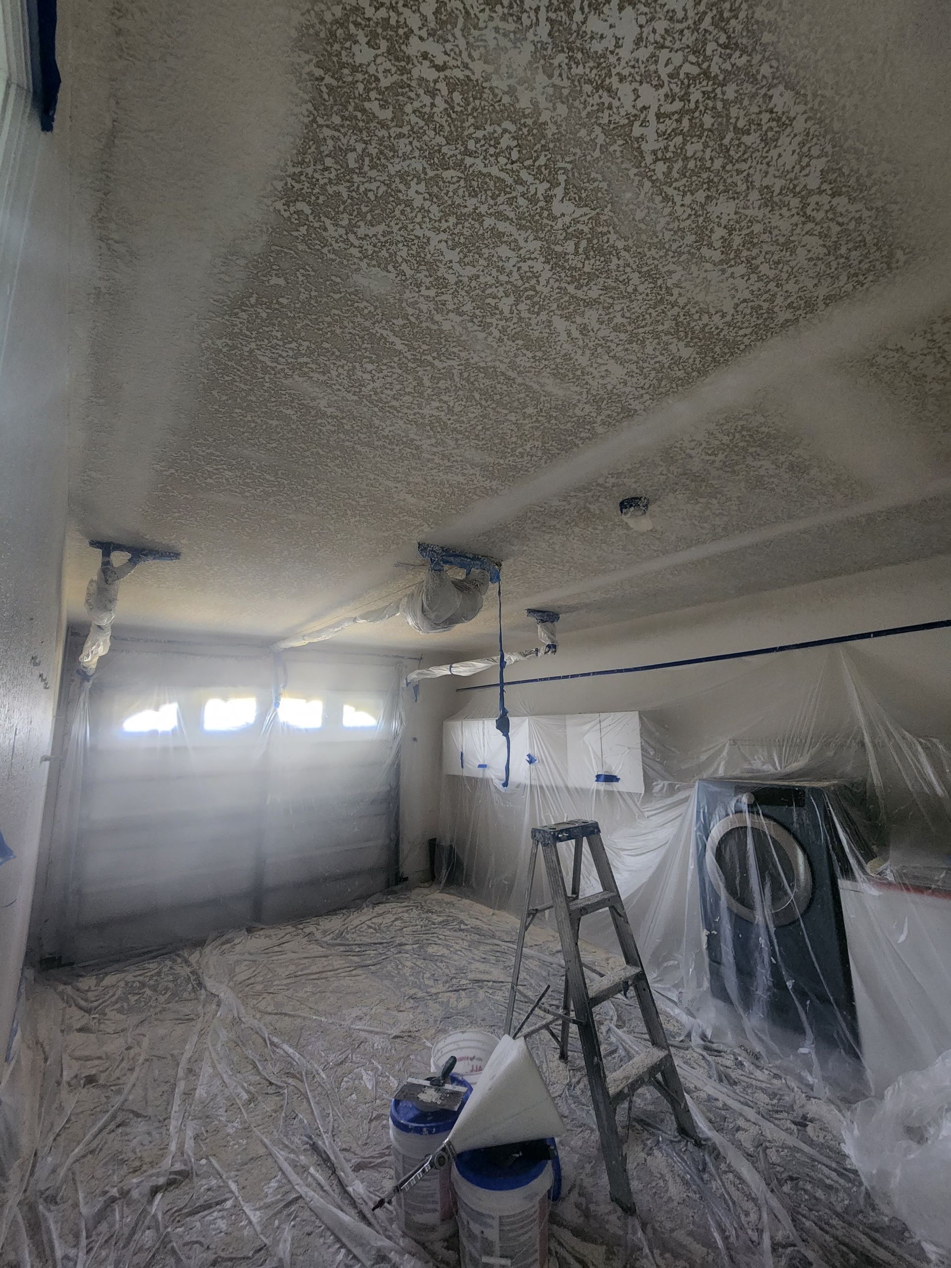 Garage under renovation, with spray foam insulation on the ceiling and walls, ladder, bucket, and covered car.