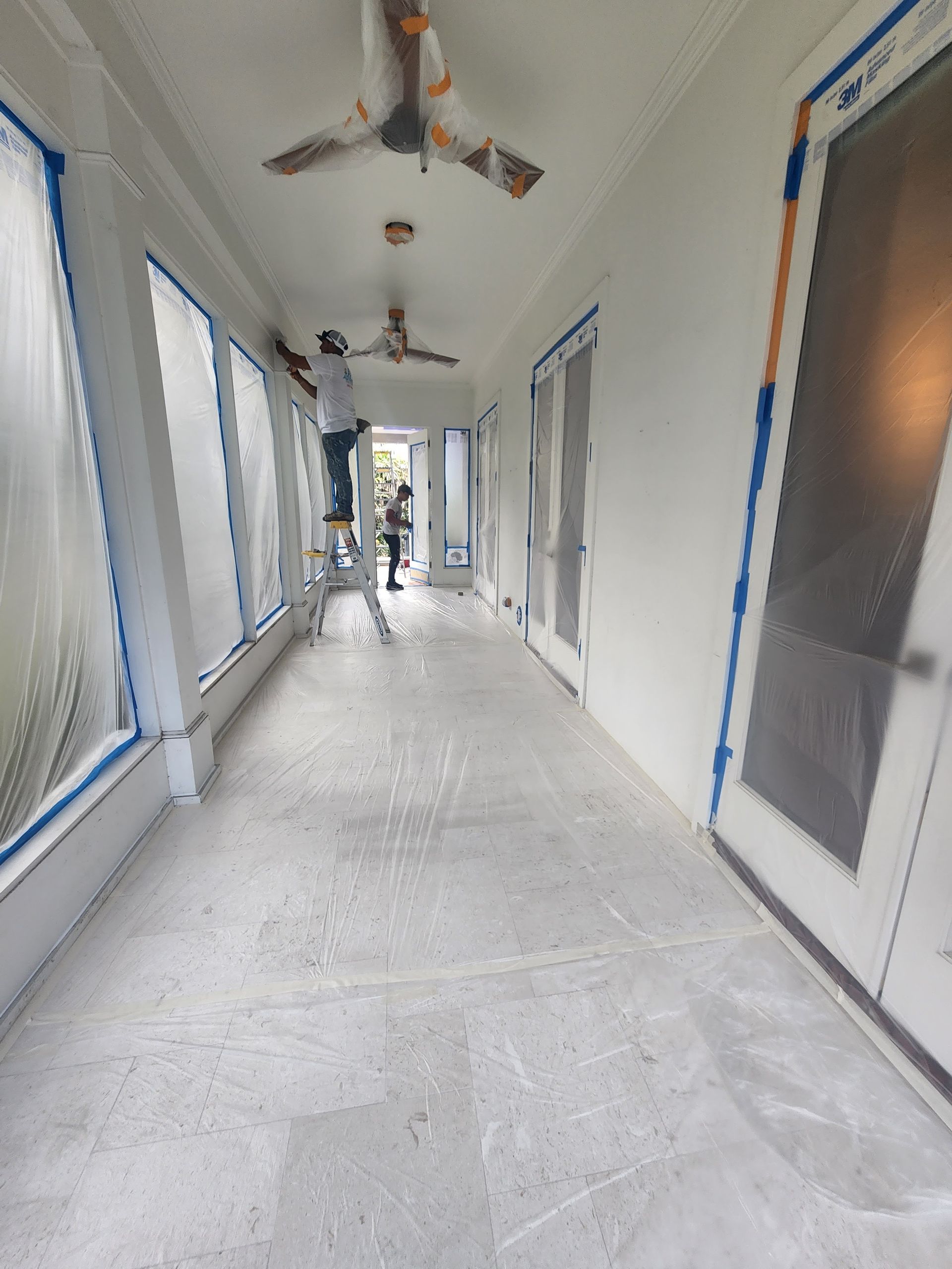 Long, unfinished hallway with white walls, blue painter’s tape, ceiling fans, and windows along the left side