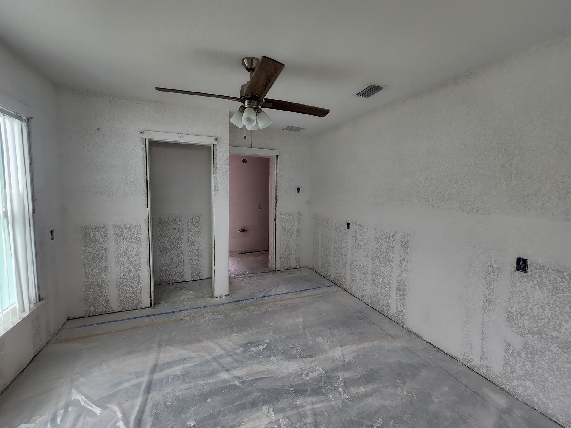 Empty white room with ceiling fan, plastic-covered floor, and doorway to a pink room