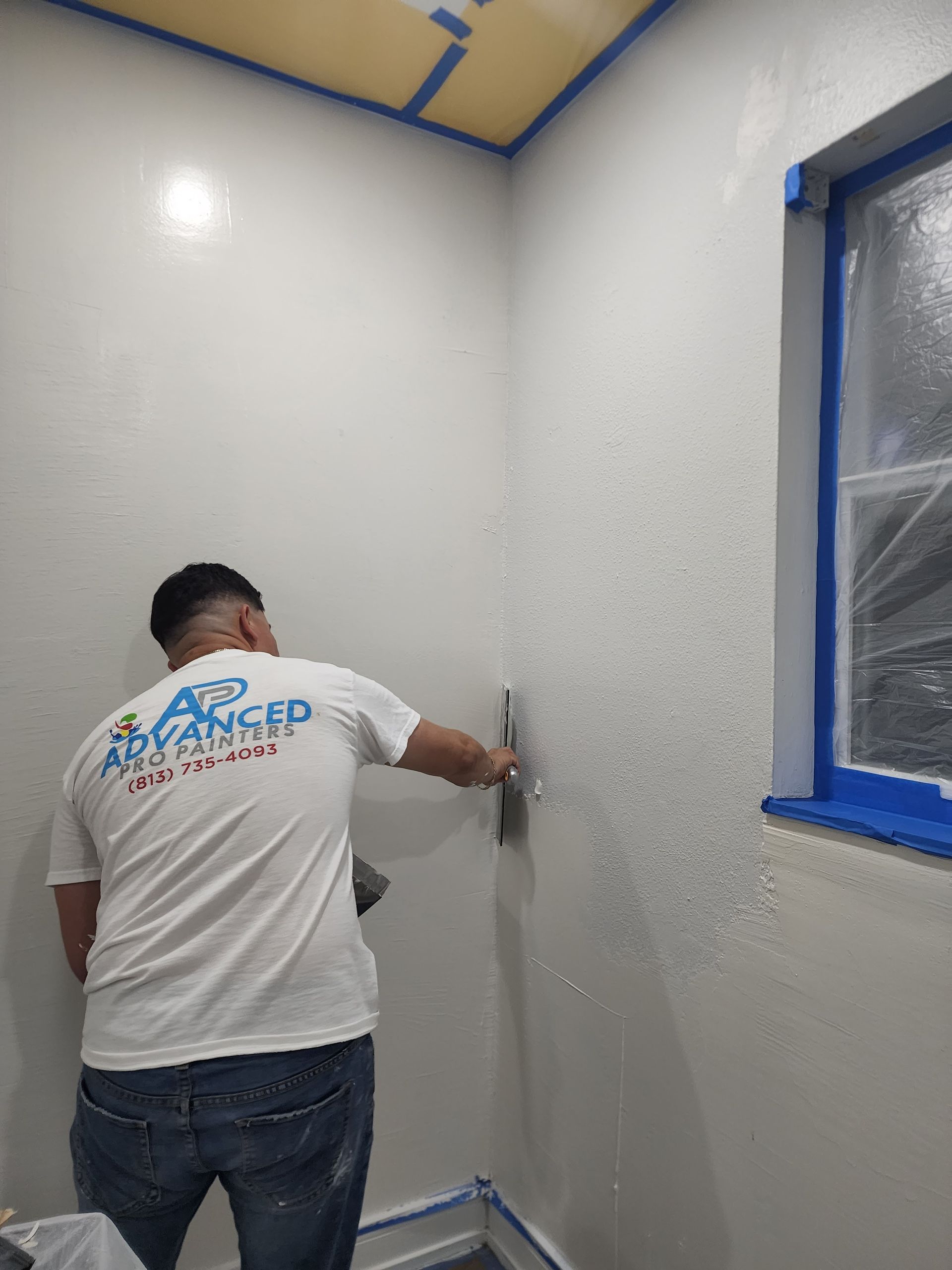 Worker painting a white corner wall inside a room, with blue tape around the window frame.