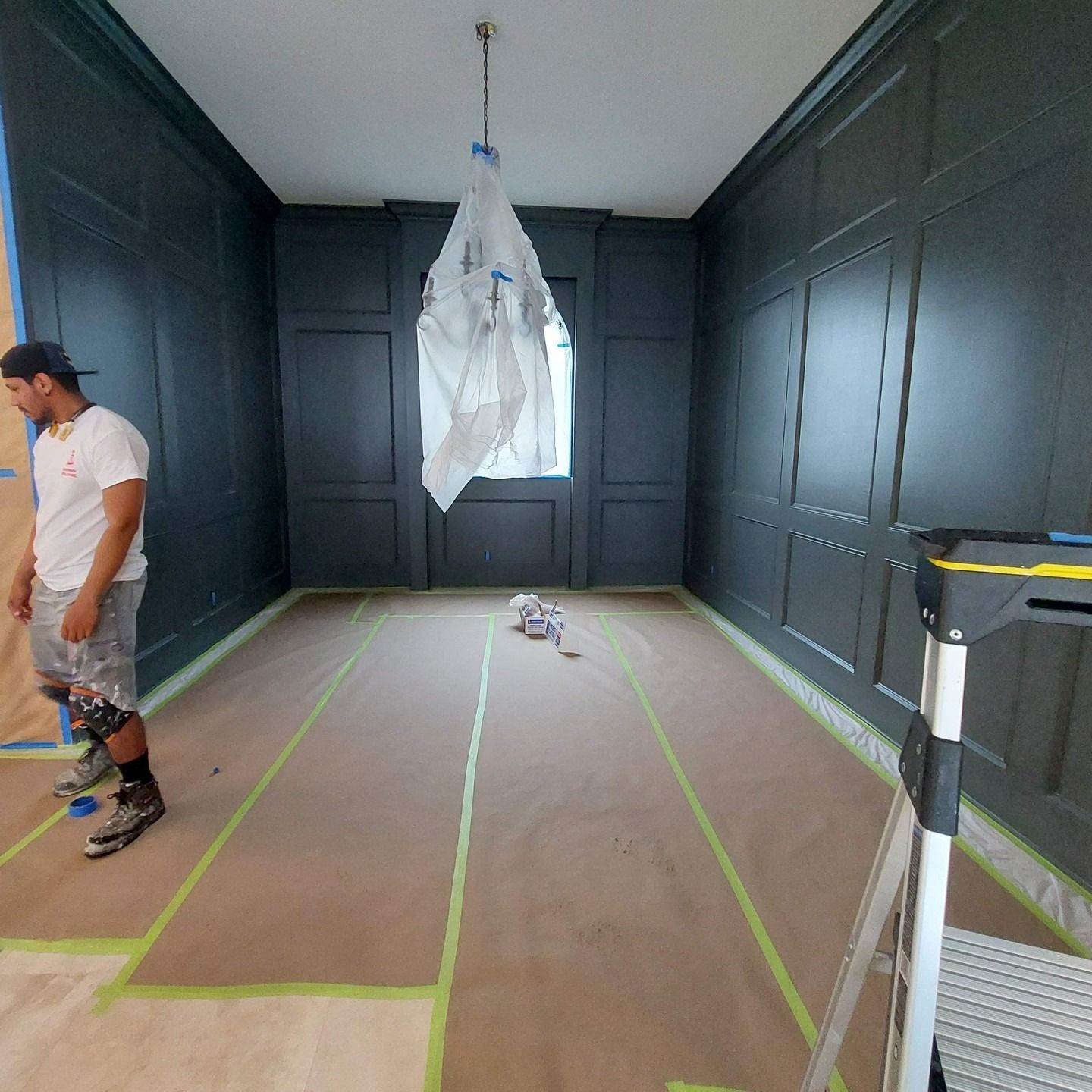 Room being painted, with dark wall panels, masked floor, a hanging plastic sheet, and a man at left.