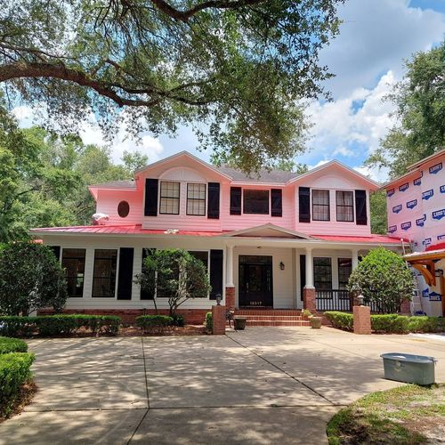Pink two-story house with a wraparound porch, black shutters, and a tree-lined driveway under a blue sky