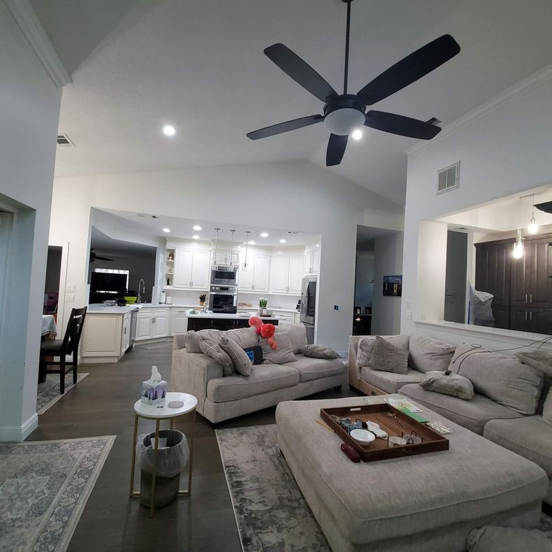 Modern open-plan living room with beige sectional sofas, ceiling fan, and bright white kitchen in the background