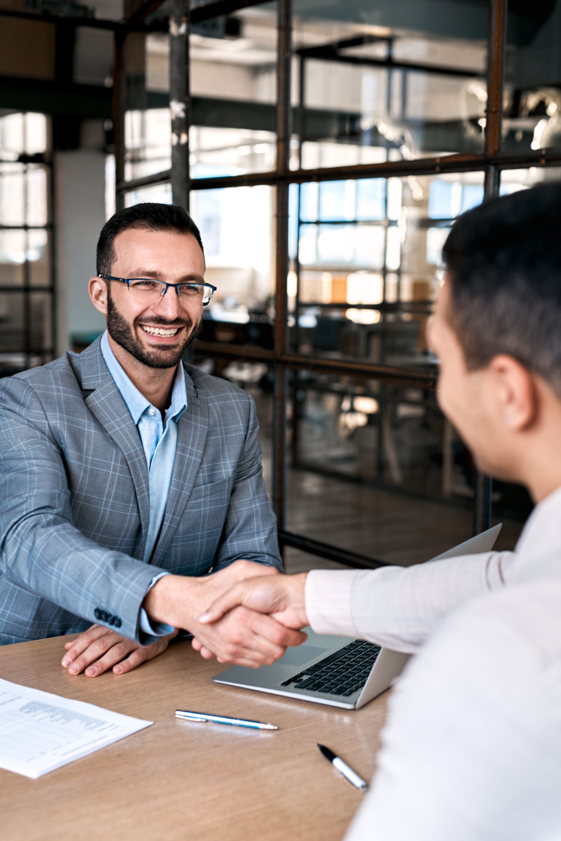 Two men shaking hands at a table, smiling. One wears a suit jacket, glasses, and a blue shirt.