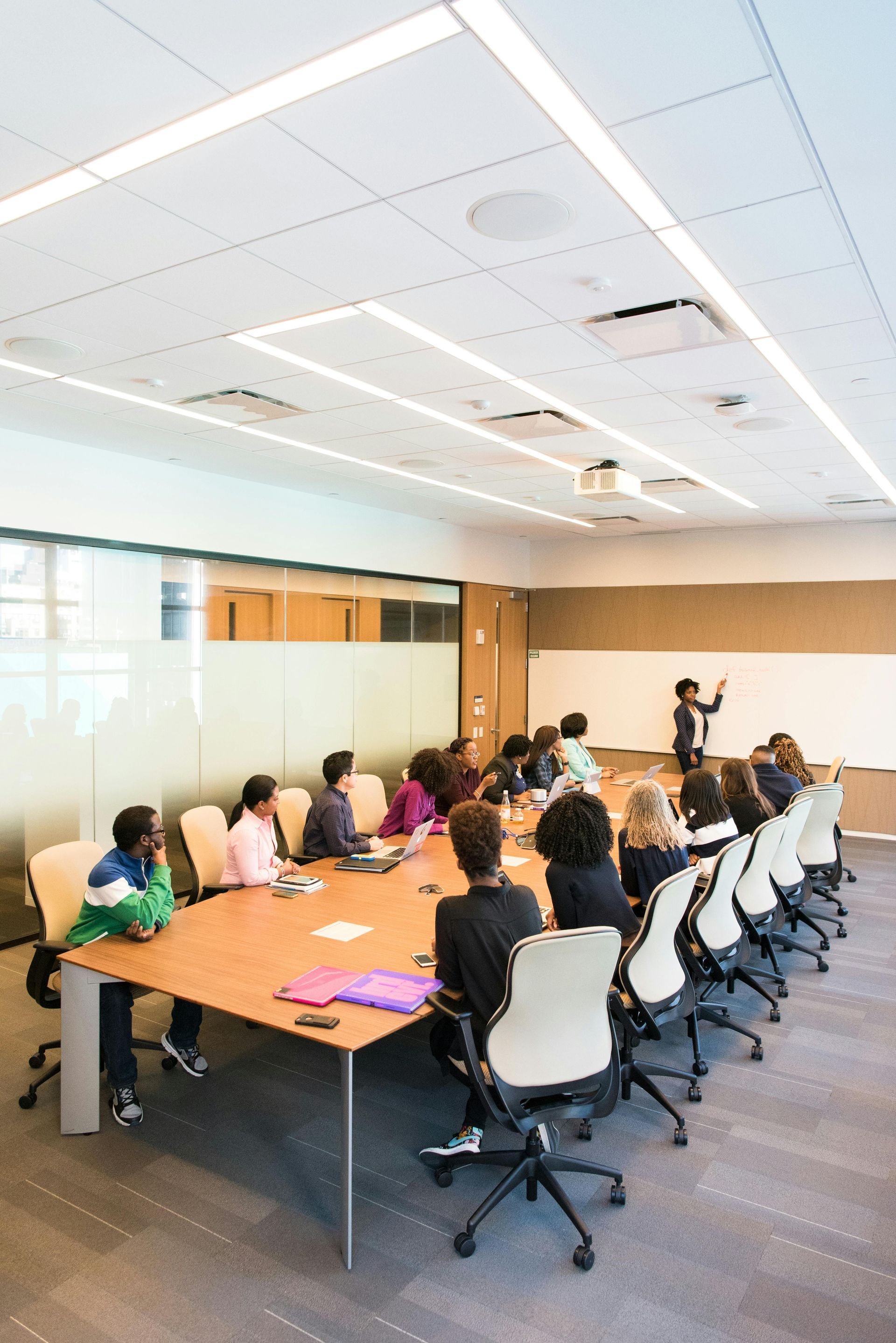 People in a meeting room, some seated around a table, one at the whiteboard. Modern office setting.