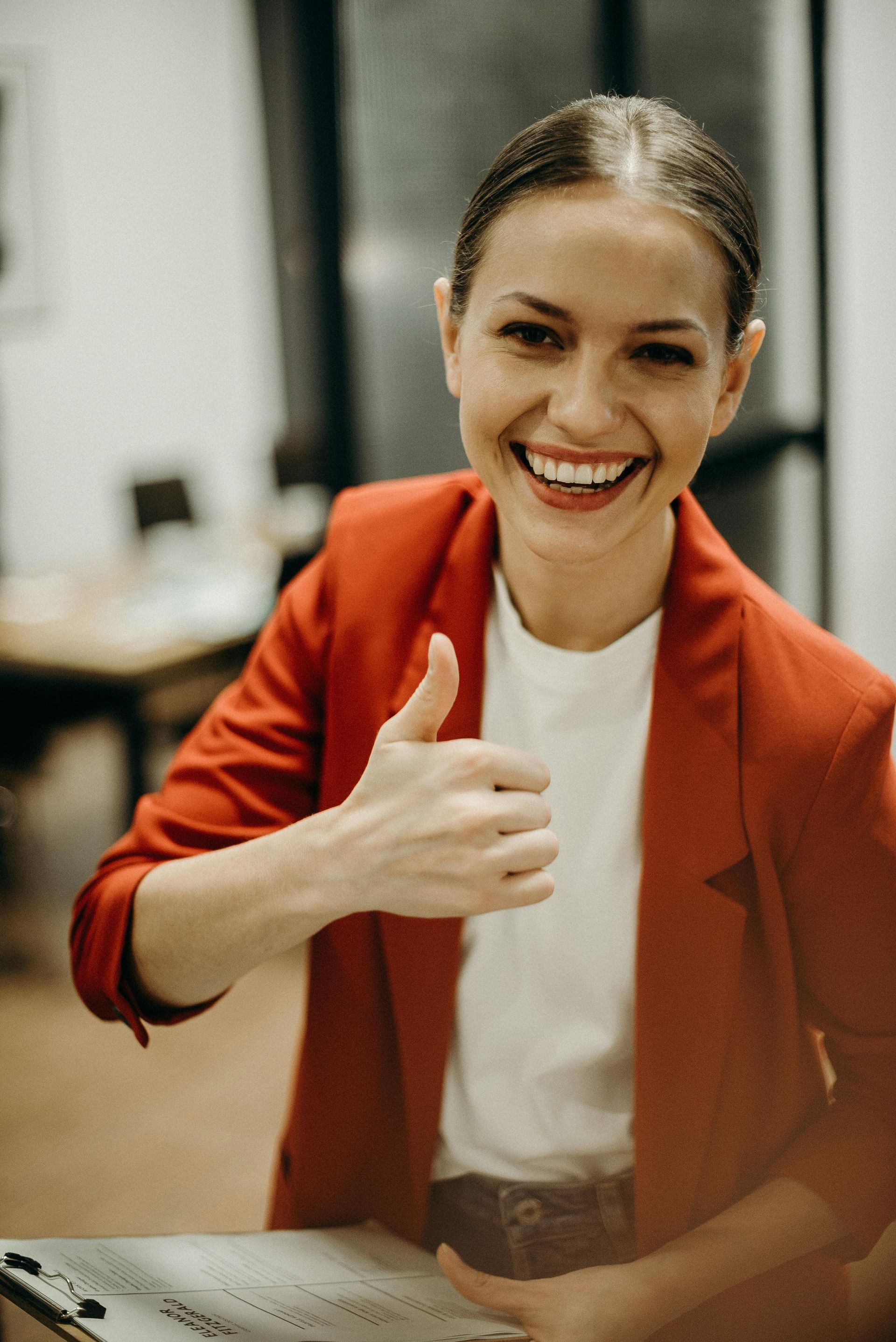 Woman in red blazer smiles and gives thumbs-up in an office setting.