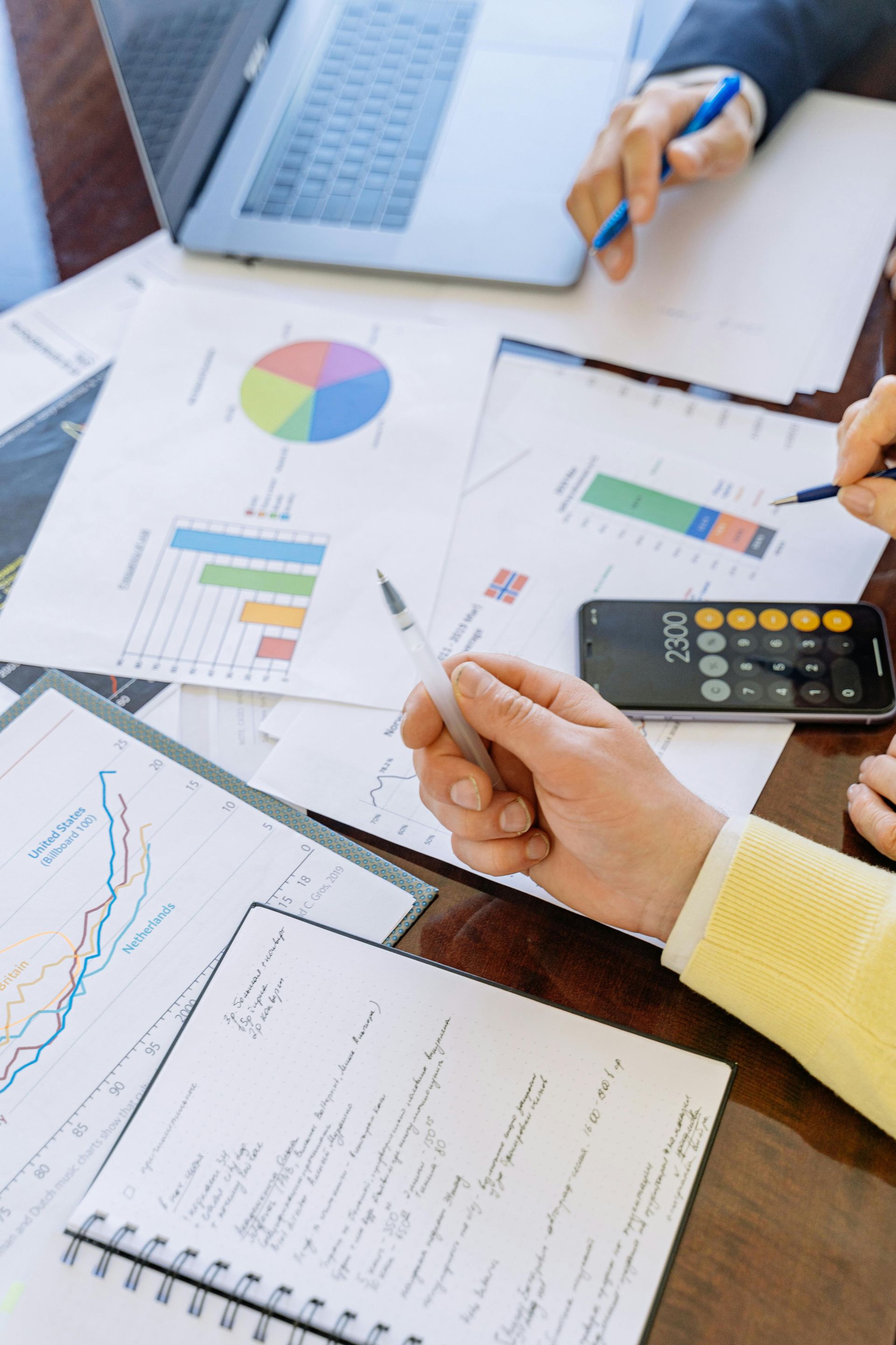 Hands analyzing financial charts and data on a table with a laptop and calculator.