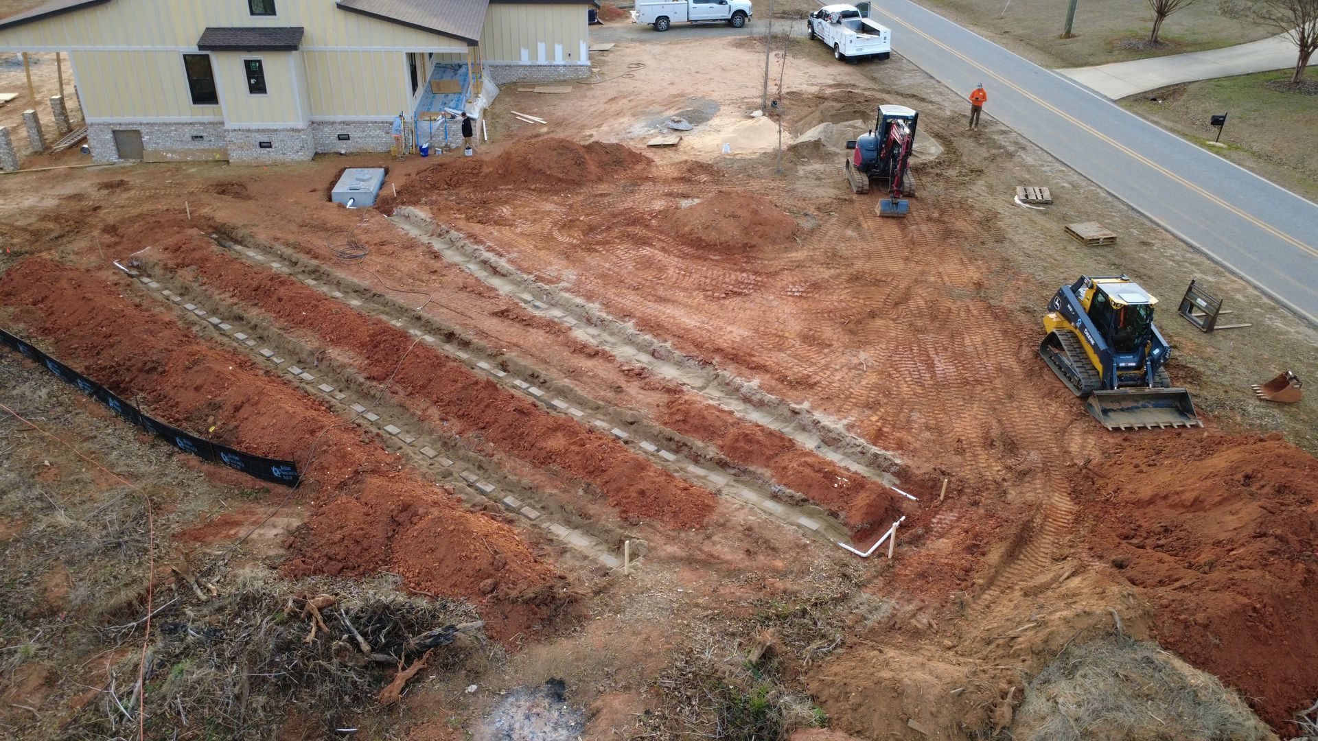 Construction site with excavators grading red dirt beside a road and house