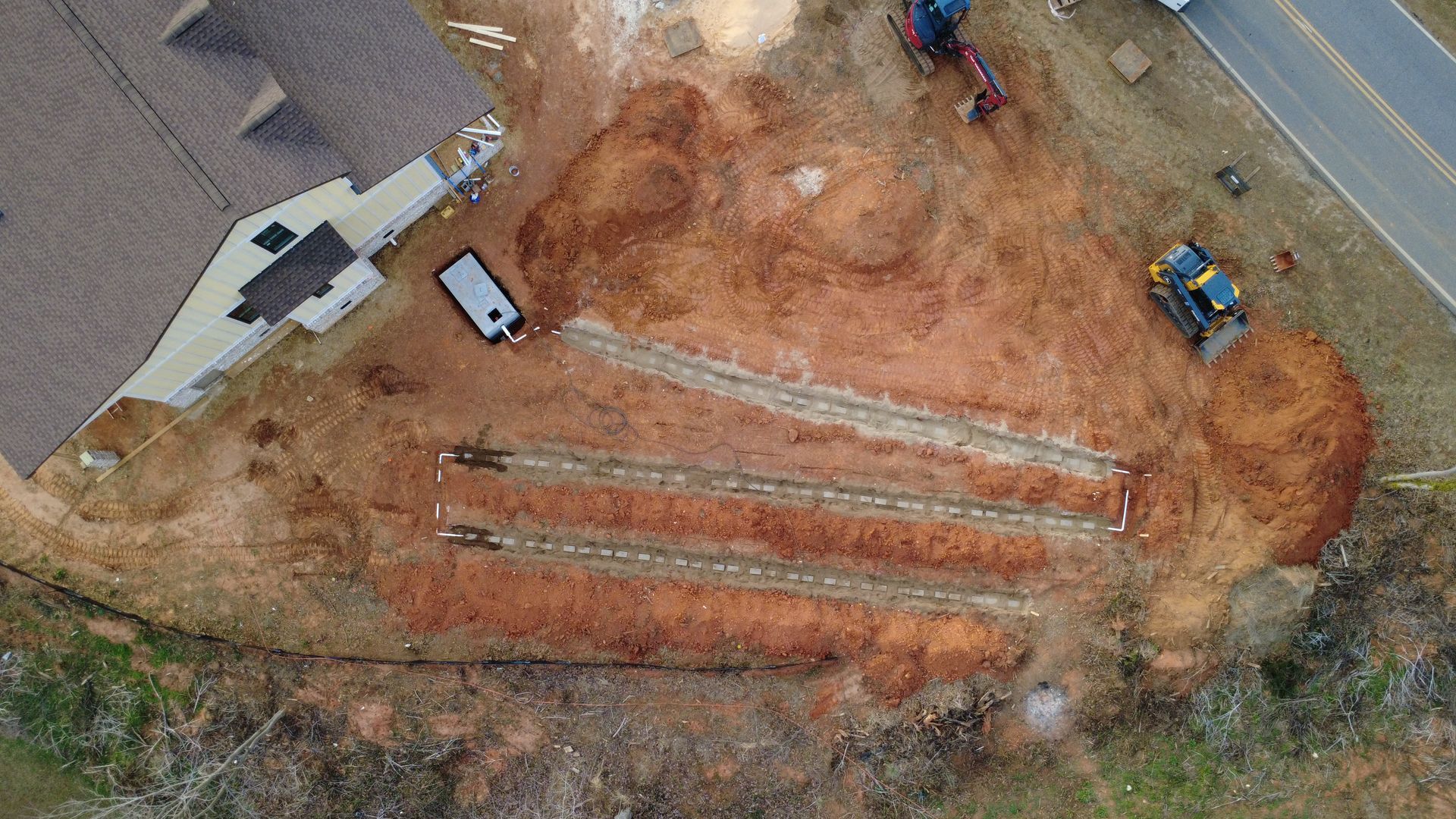 Aerial view of a construction site with three long trenches, dirt piles, and vehicles on a cleared lot