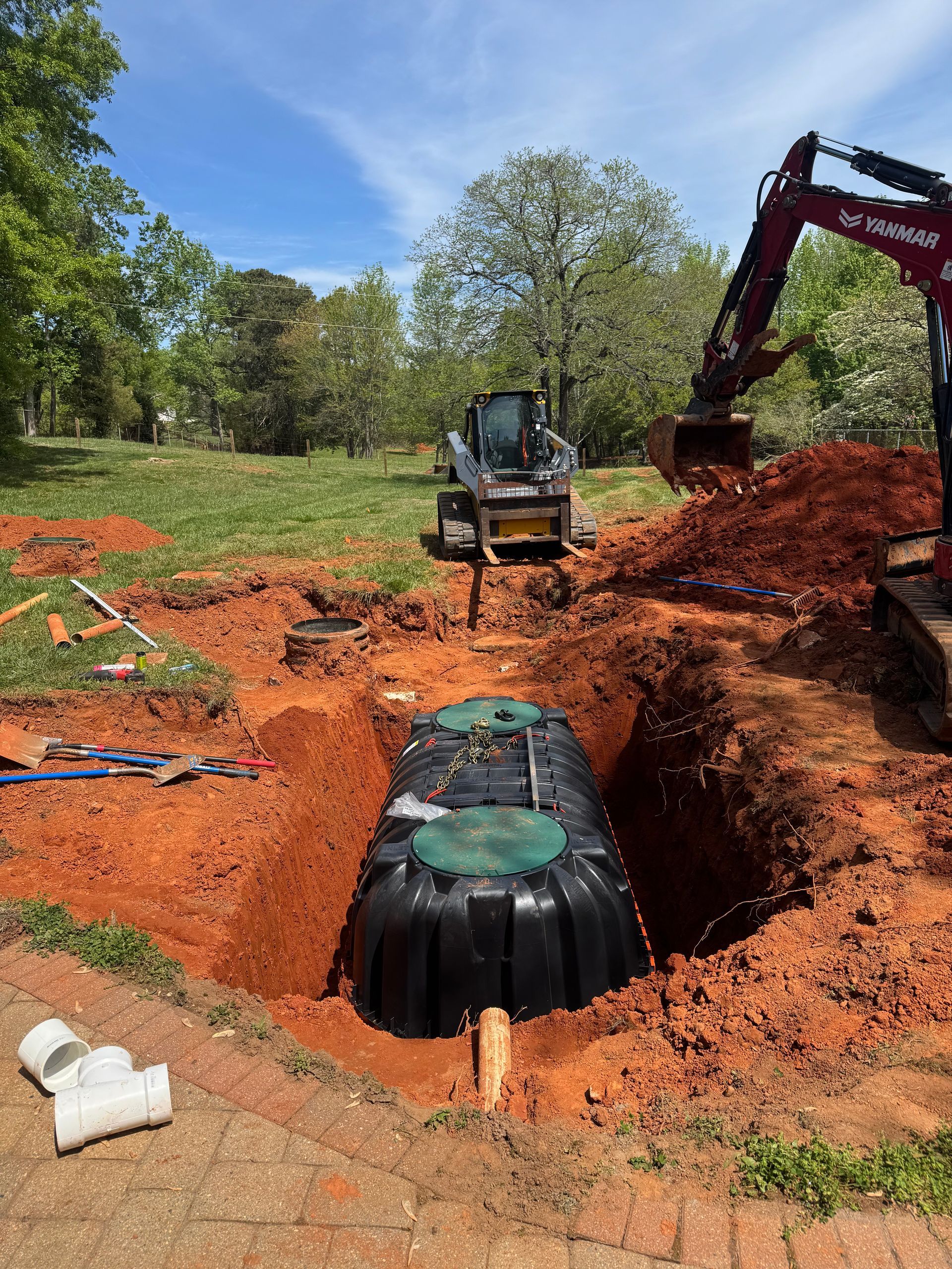 Excavator installing a large black tank in a red clay trench on a grassy construction site