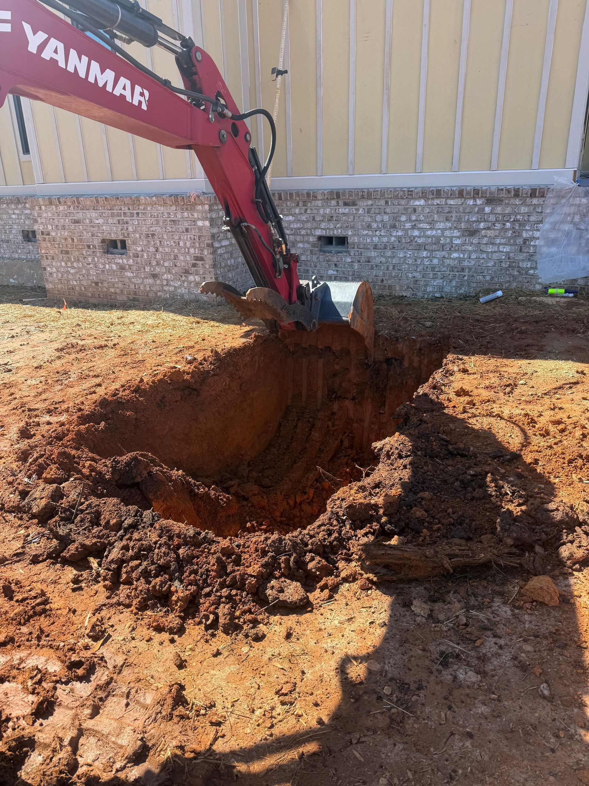 Red excavator digging a large hole beside a building wall in muddy soil