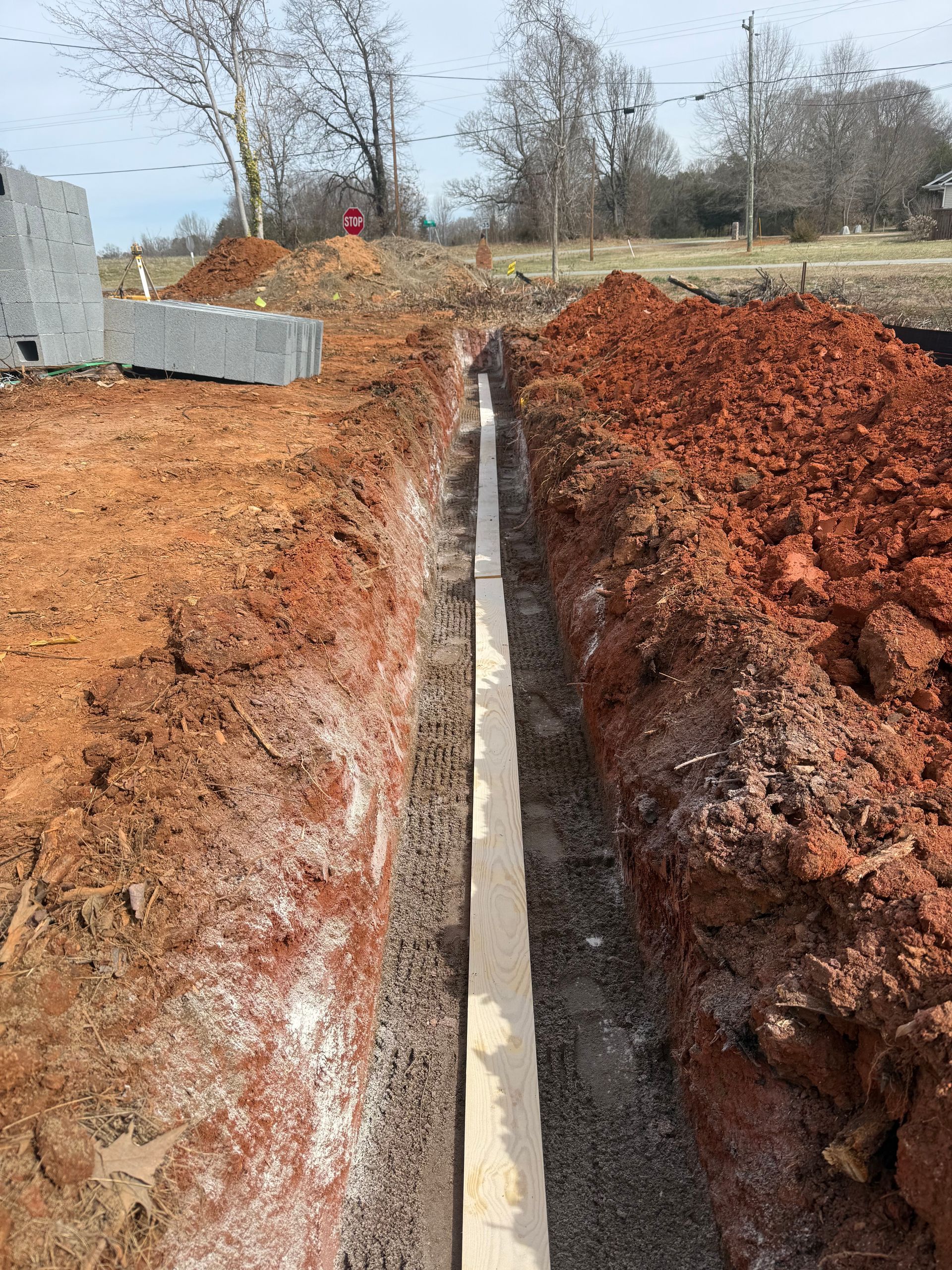 Deep trench cut through red soil at a construction site, with concrete pipe and equipment nearby.