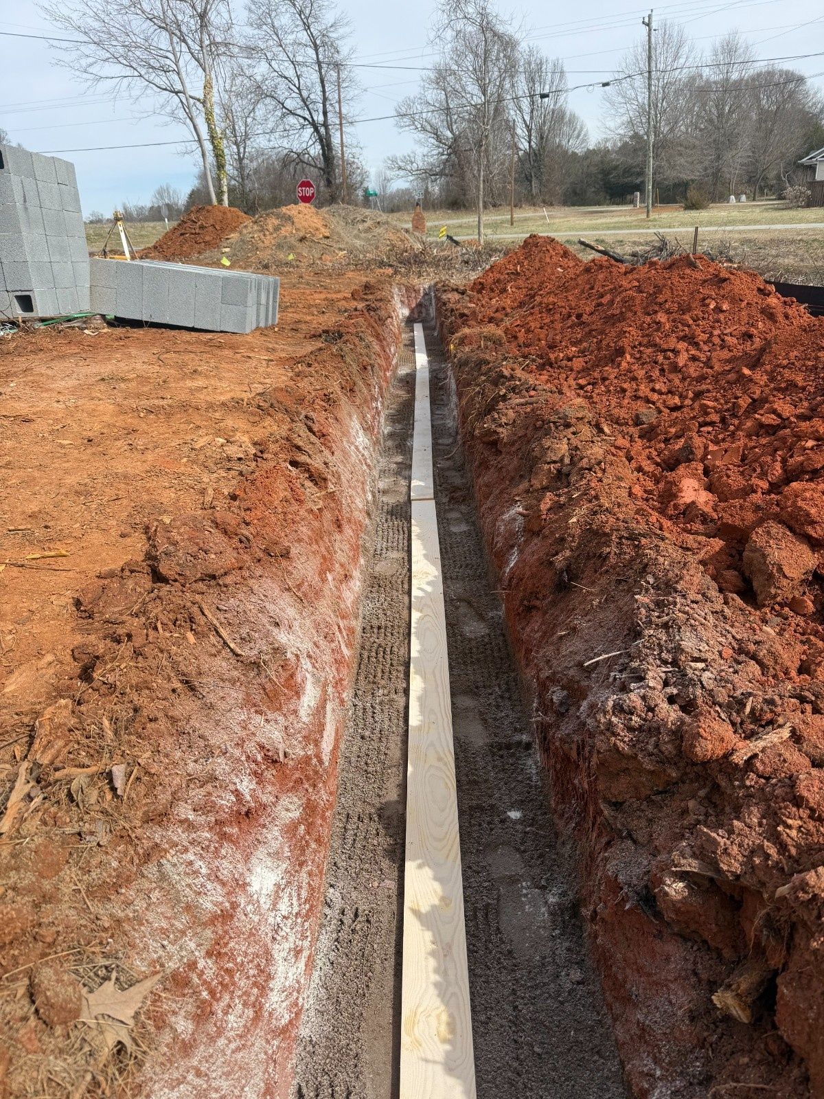 Narrow trench with concrete footing between piles of red dirt at a construction site
