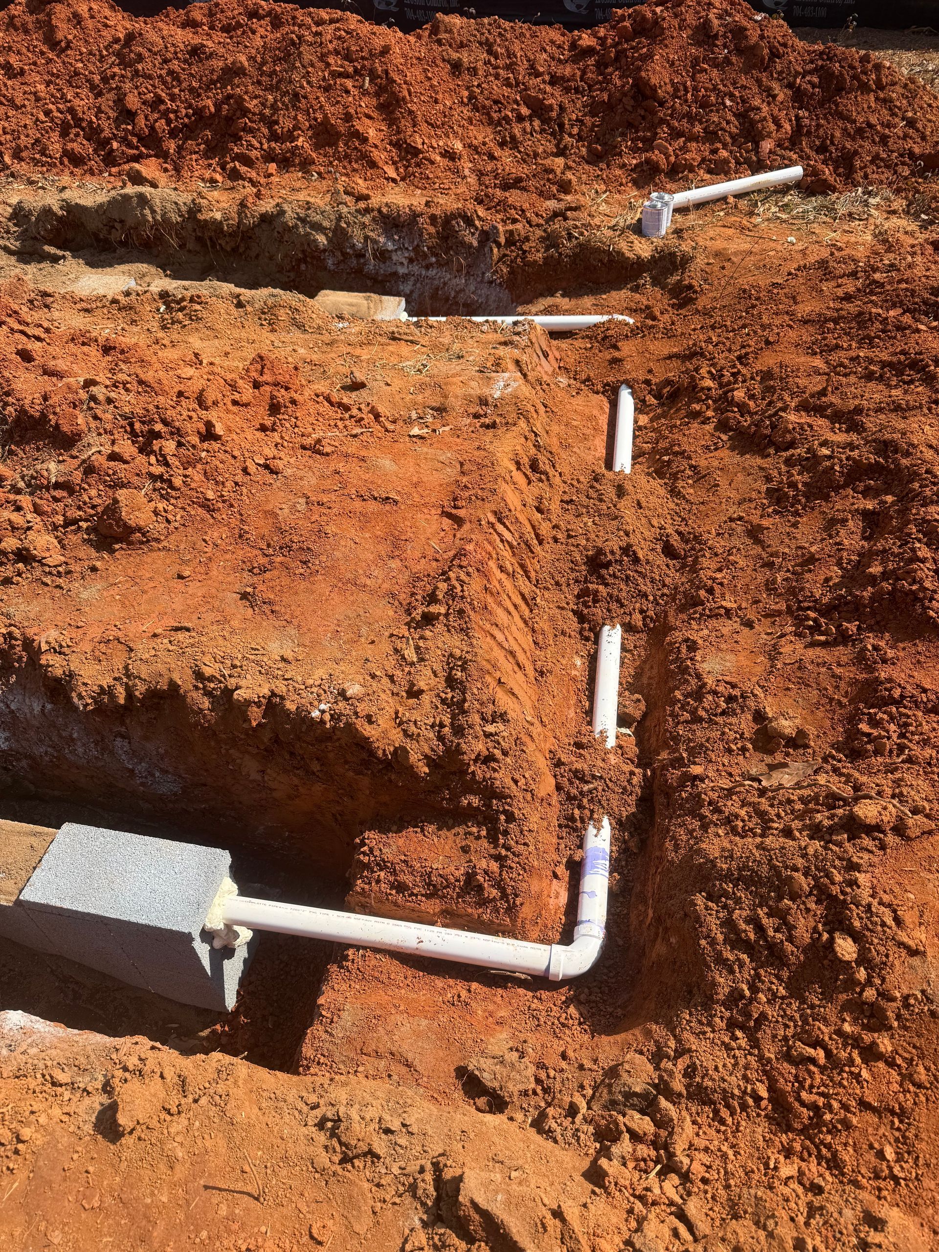 White PVC pipe and elbow joint installed in a reddish dirt trench, with a concrete block nearby.