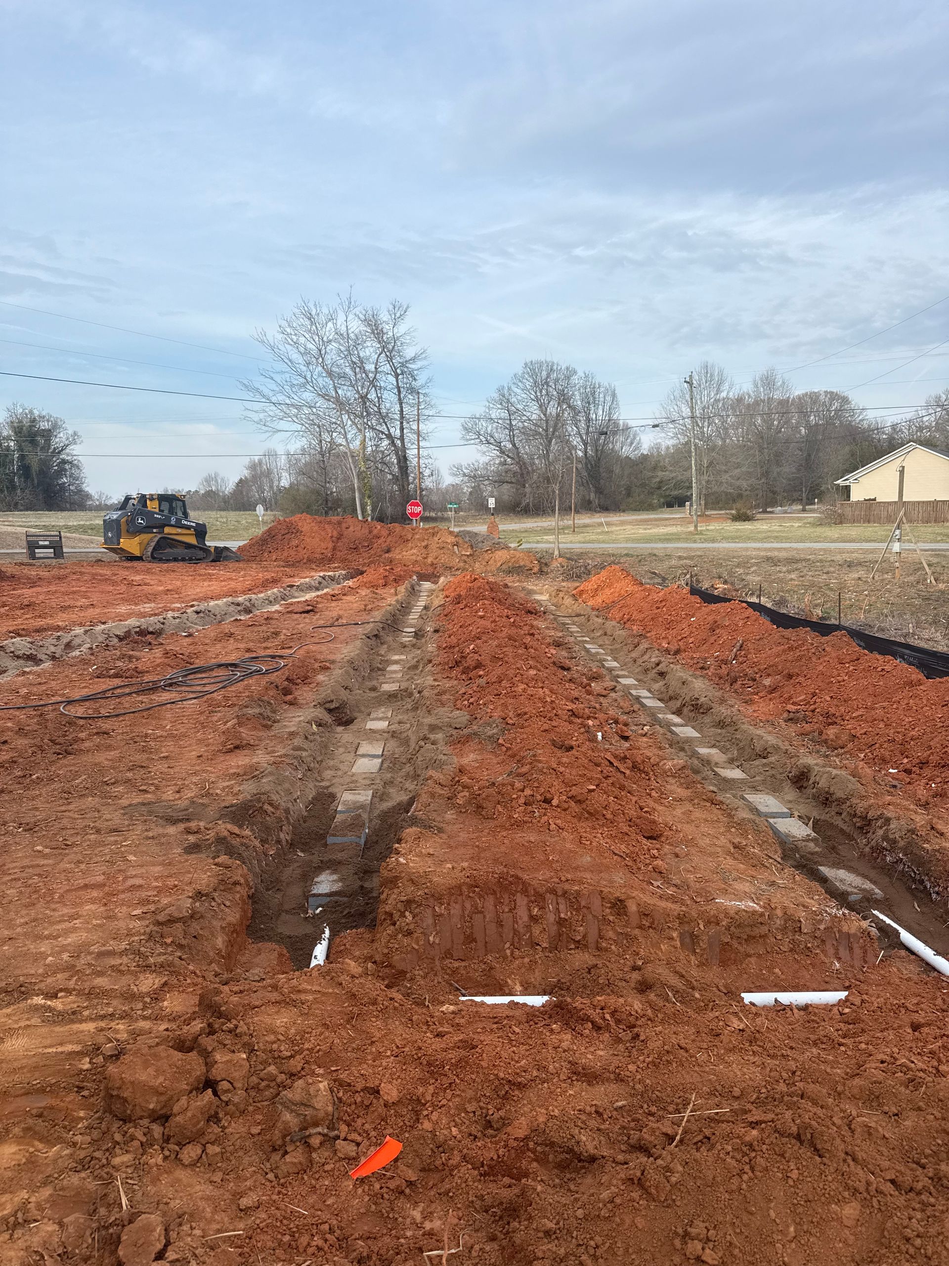 Mud-filled construction site with deep tire tracks, a yellow excavator, and bare trees under a cloudy sky